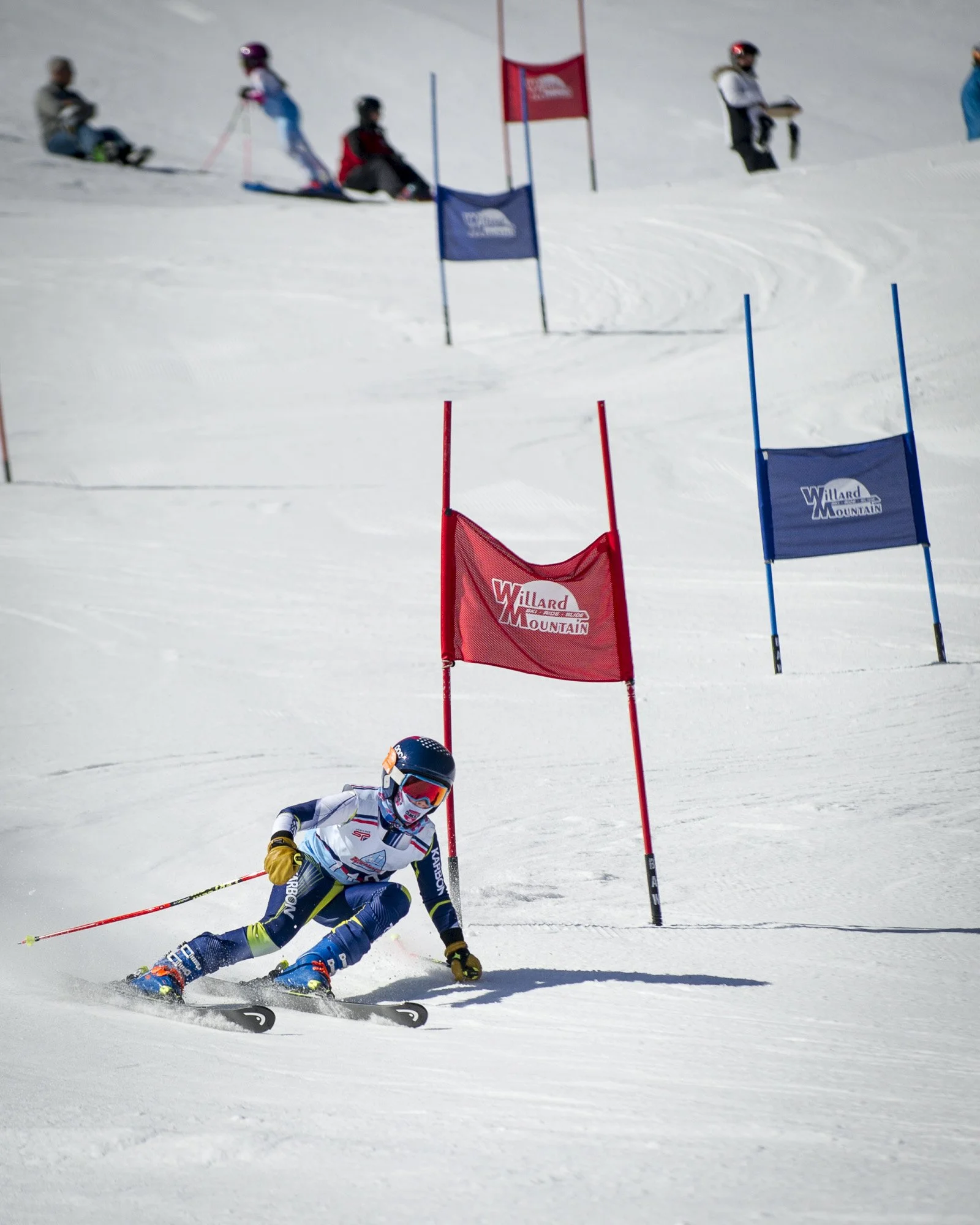 A young skier in racing gear navigating a slalom course on snow, passing red and blue gates with other children and an adult in the background.