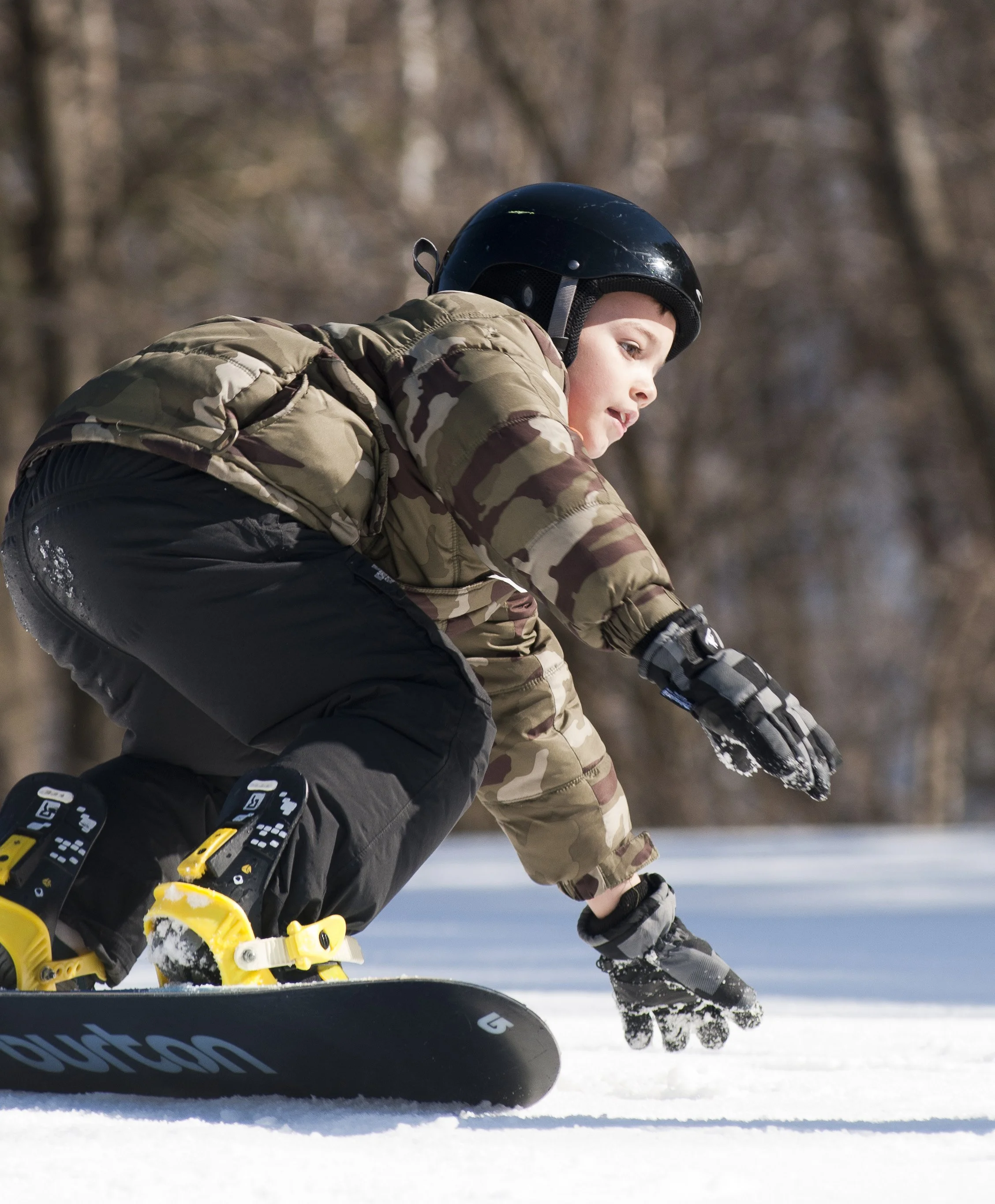 A young boy wearing a black helmet, camouflage jacket, and gloves snowboarding on snow with a wooded background.