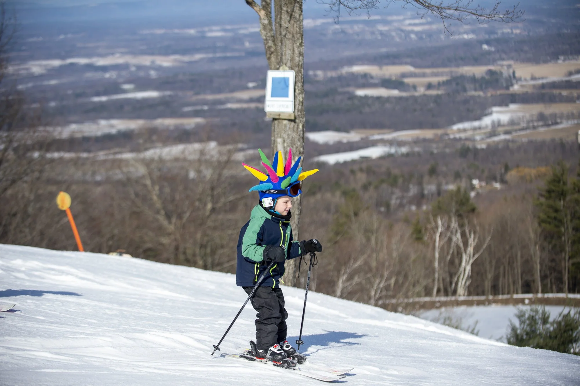 A young child skiing down a snowy slope, wearing a colorful, spiky hat resembling a cartoon character, a blue and green winter jacket, black ski pants, gloves, and goggles.