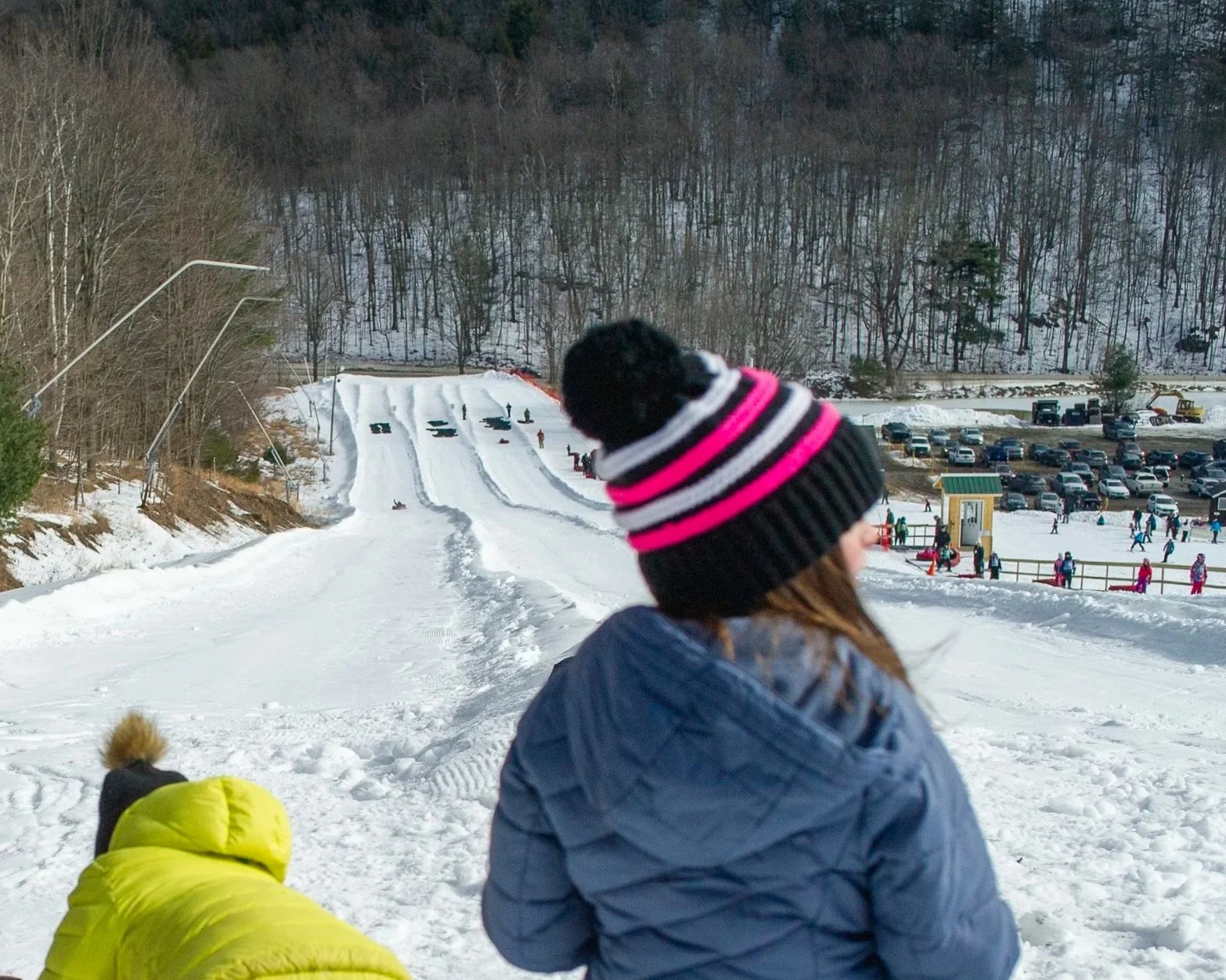 A child with a black, white, and pink striped knit hat looks towards a snow-covered ski slope with other people skiing and snowboarding. There are vehicles and a parking lot in the background, with a wooded hill.