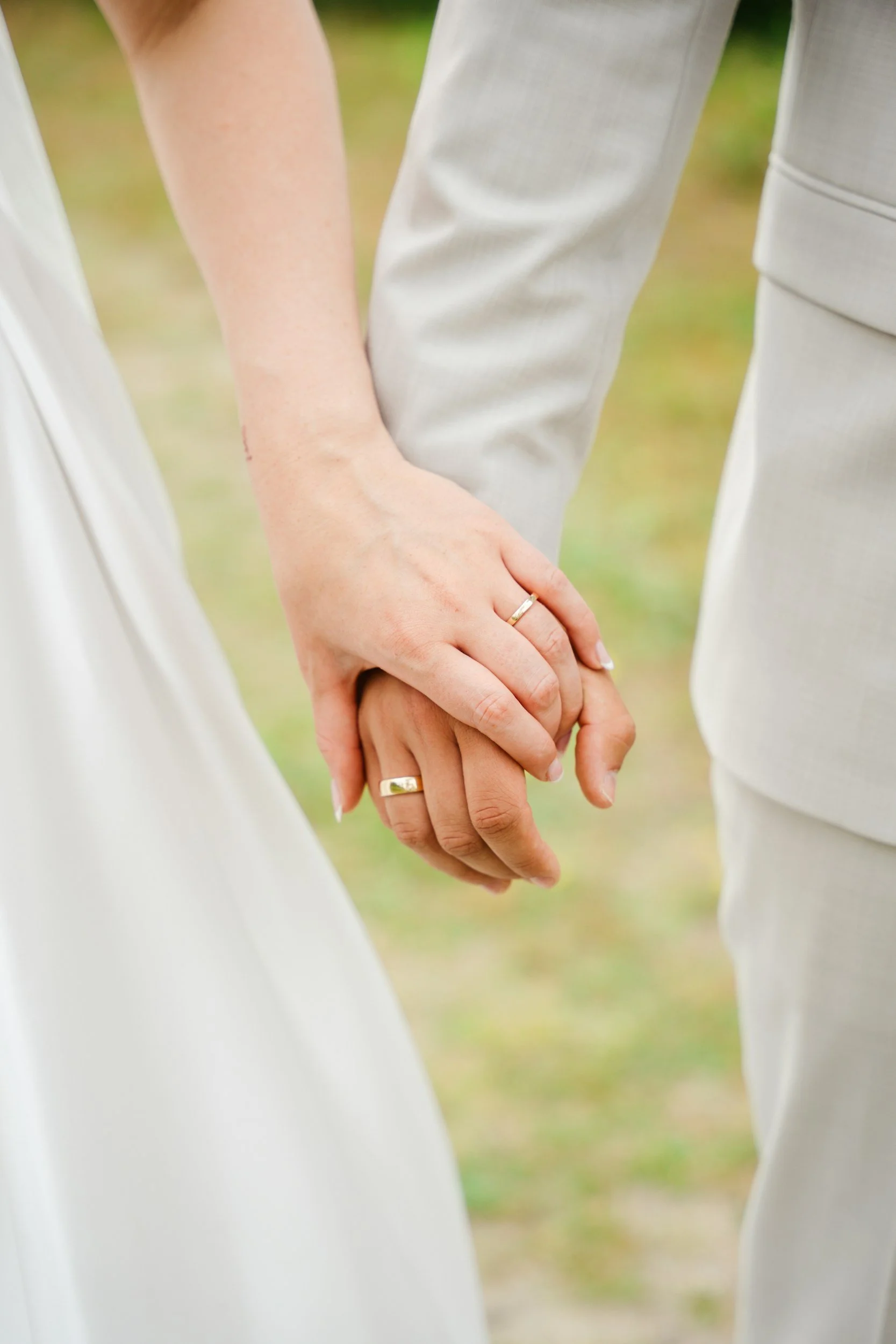 Duas pessoas segurando as mãos uma da outra, usando roupas brancas e anéis de casamento.