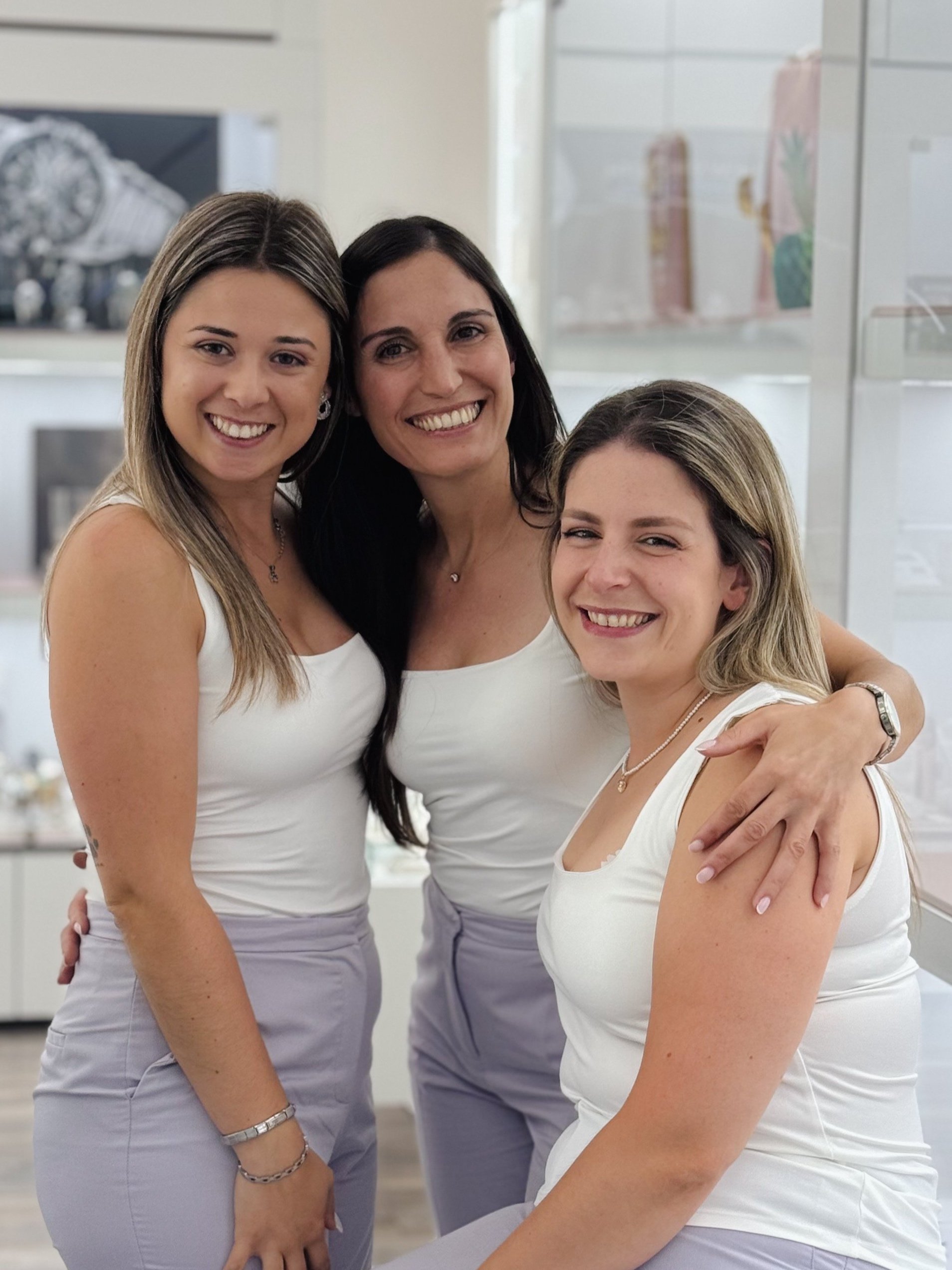 Três mulheres sorrindo, vestidas com roupas brancas e em um ambiente de ourivesaria.