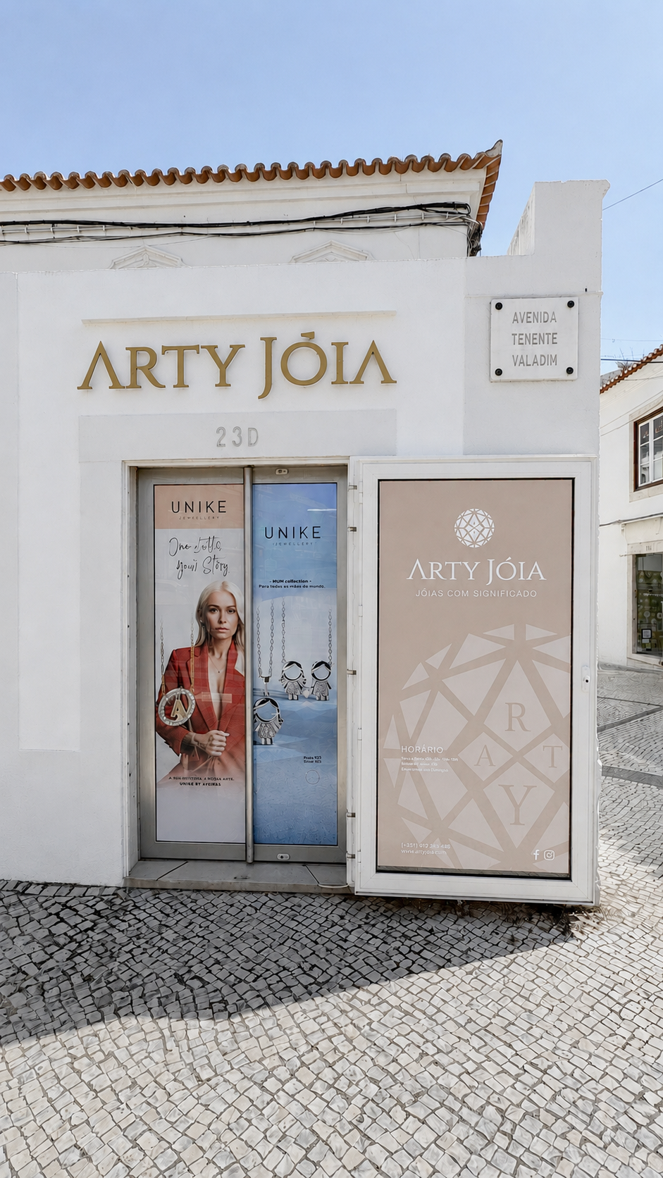 Fachada de uma loja de joias chamada Arty Jóia, com letreiro dourado, porta de vidro com anúncios de joias e publicidade com uma mulher usando joias, na rua de calçada de pedras em uma cidade brasileira.