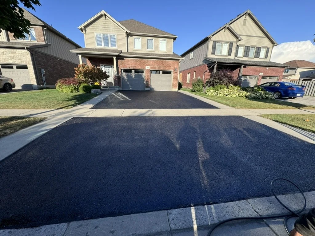 A newly paved asphalt driveway leading to two attached houses with garages, surrounded by front lawns, shrubs, and plants, under a clear blue sky.