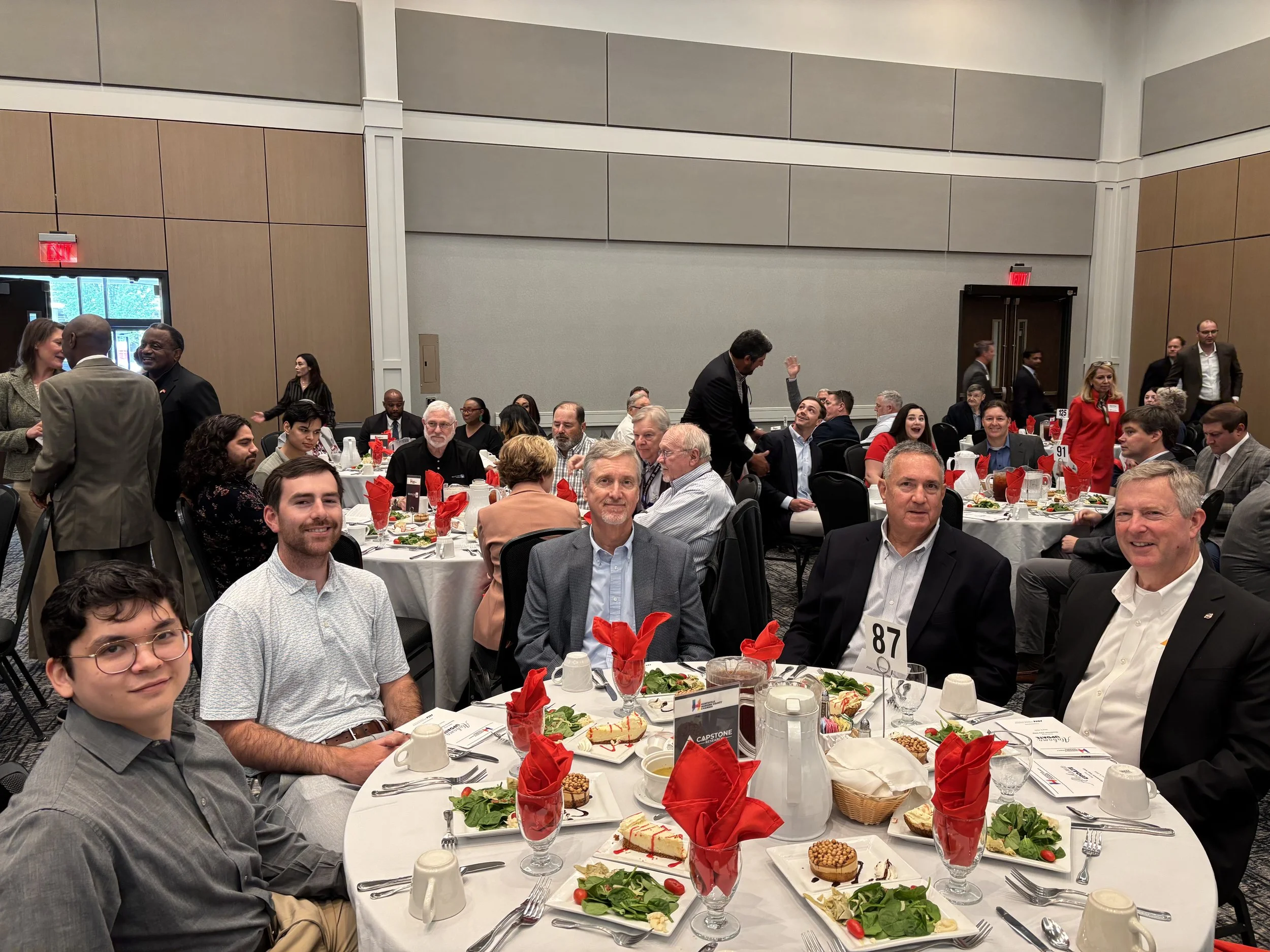 Group of people seated at round banquet tables with festive red napkins, salads, and plates of food during a formal event or banquet.