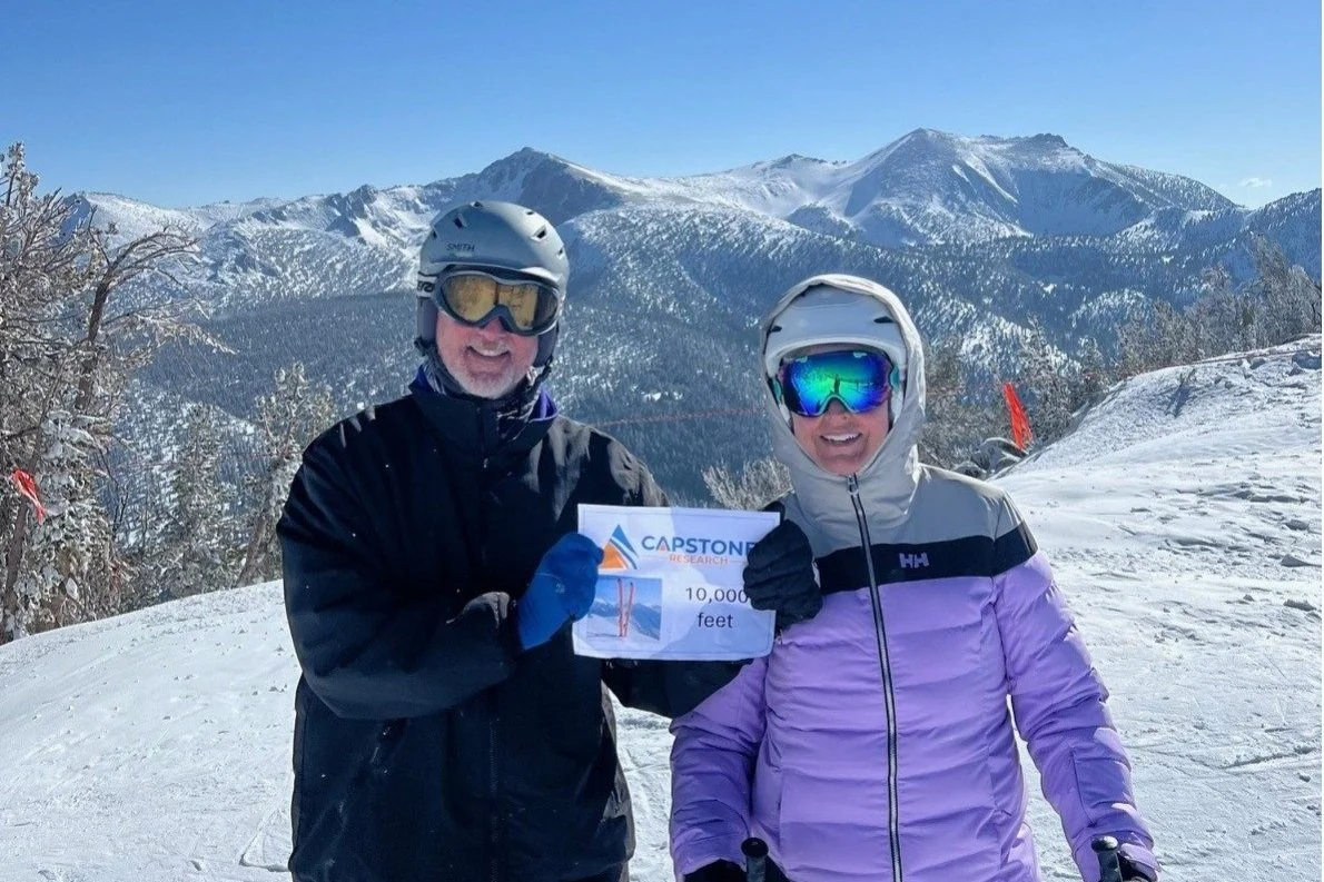 Two people in winter gear, wearing helmets and goggles, standing on a snowy mountain with snow-covered trees and mountain peaks in the background. They are holding a sign that reads "Capstone Research, 10,000 feet."