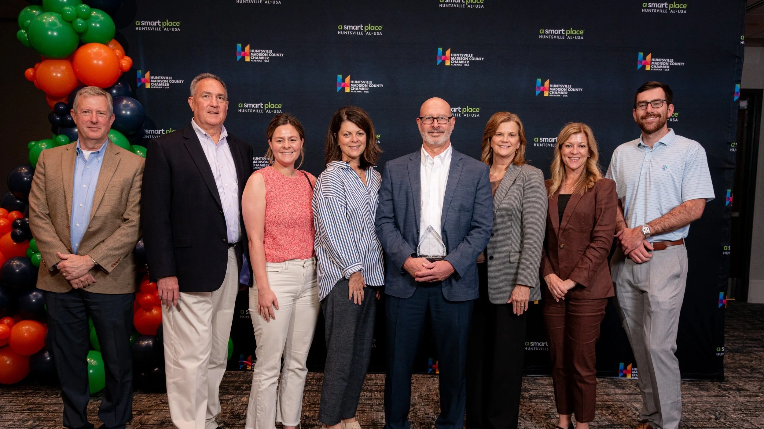 A group of eight people standing side by side at an event, with a dark backdrop featuring logos for 'a smart place' and 'Huntsville Madison County Chamber' and balloons in green, orange, and black on the left.