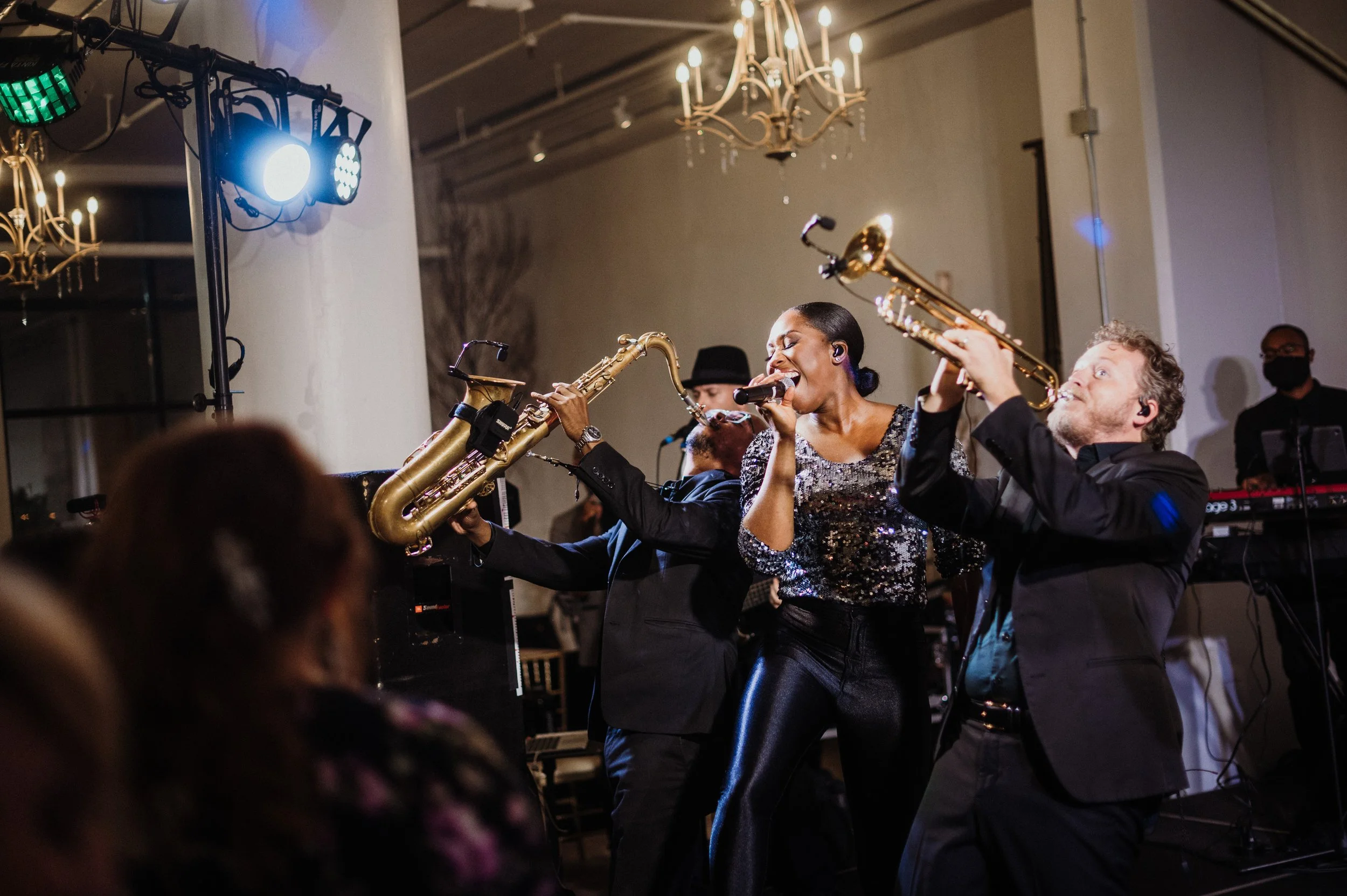 Musicians performing with saxophones and microphone at a lively event, audience in foreground, stage lights overhead.