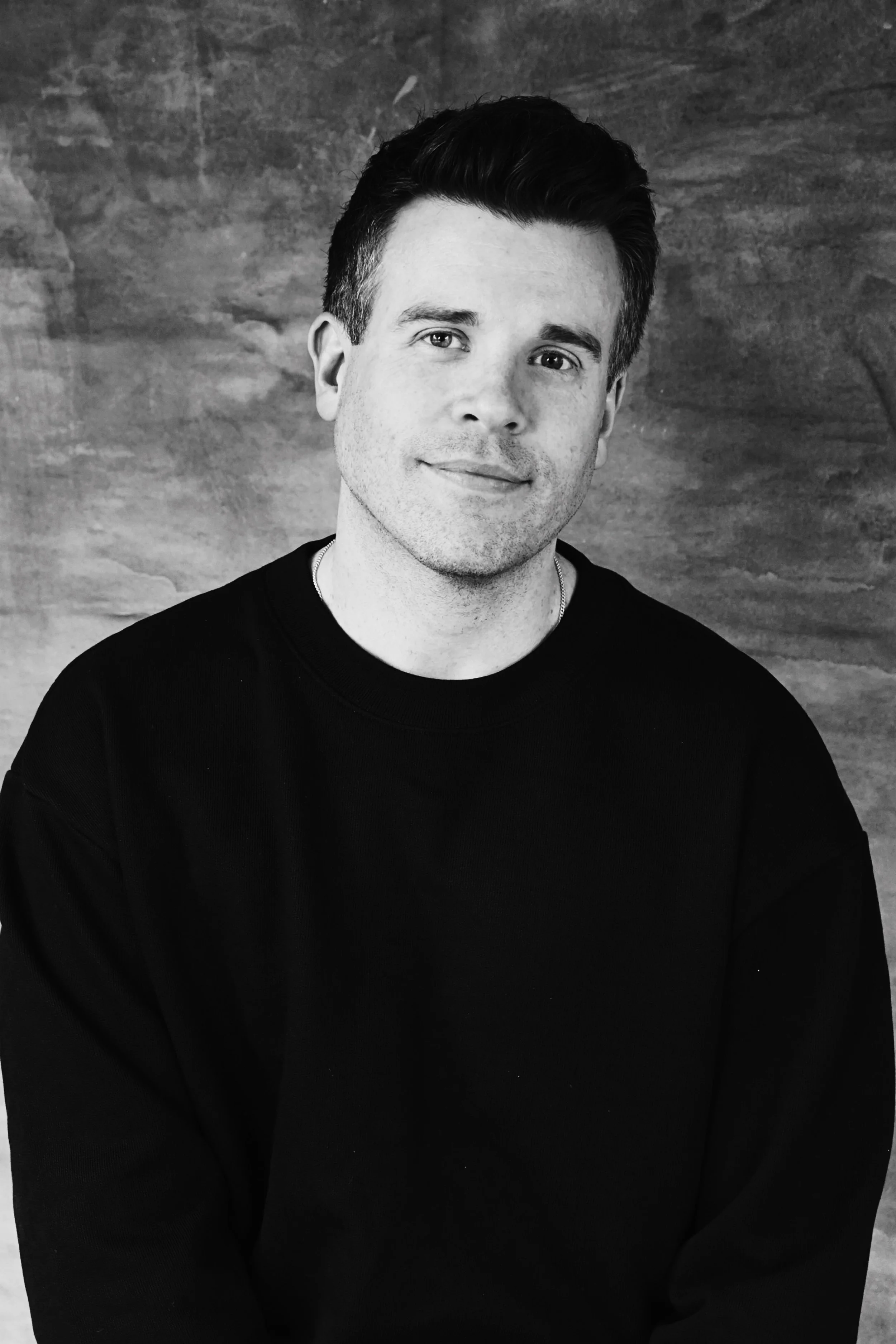A black and white portrait of a young man with short dark hair, wearing a black long-sleeve shirt, smiling softly against a wood-textured background.