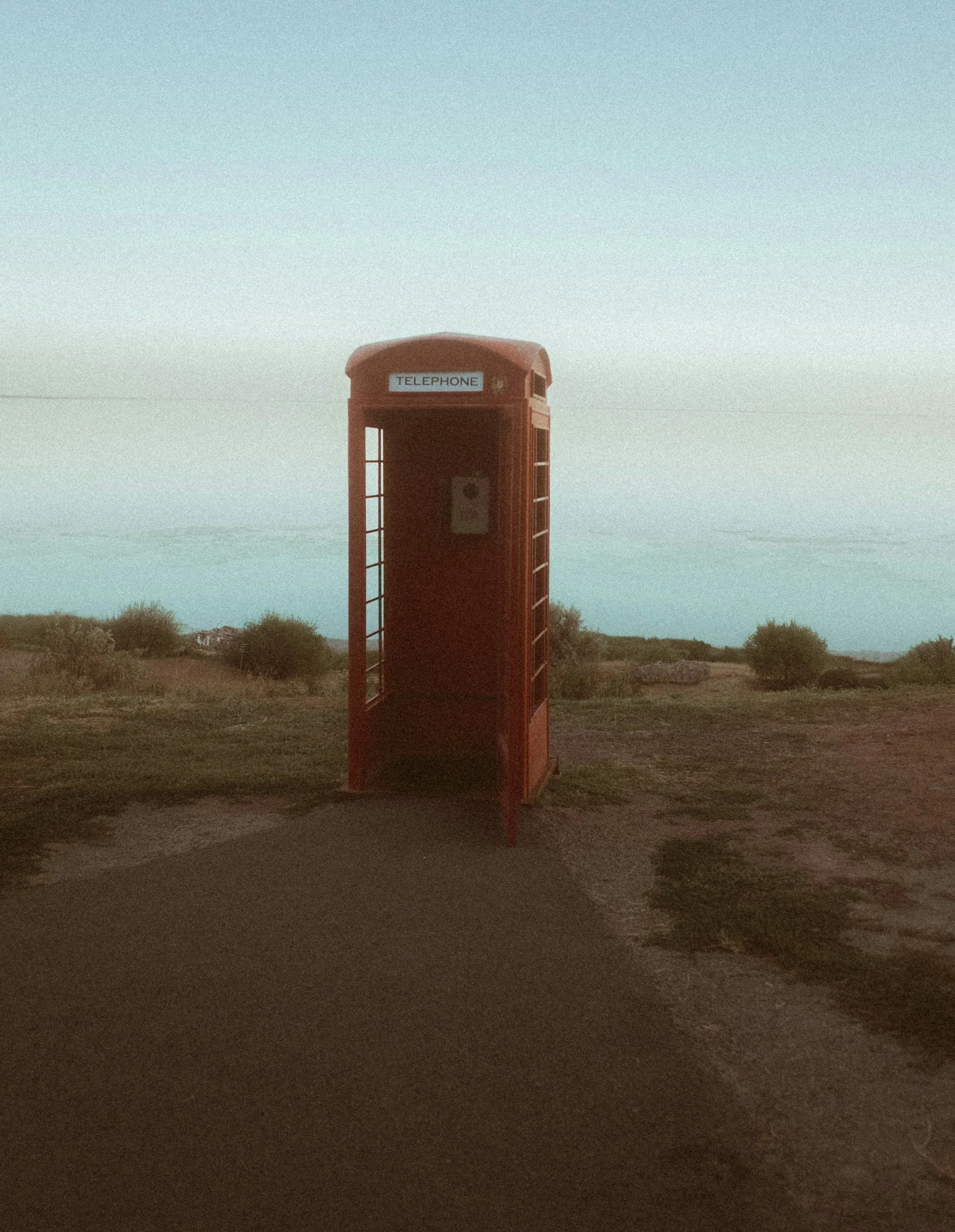 A classic red British telephone booth standing alone on a rural dirt path with sparse bushes in the distance under a mostly clear sky.