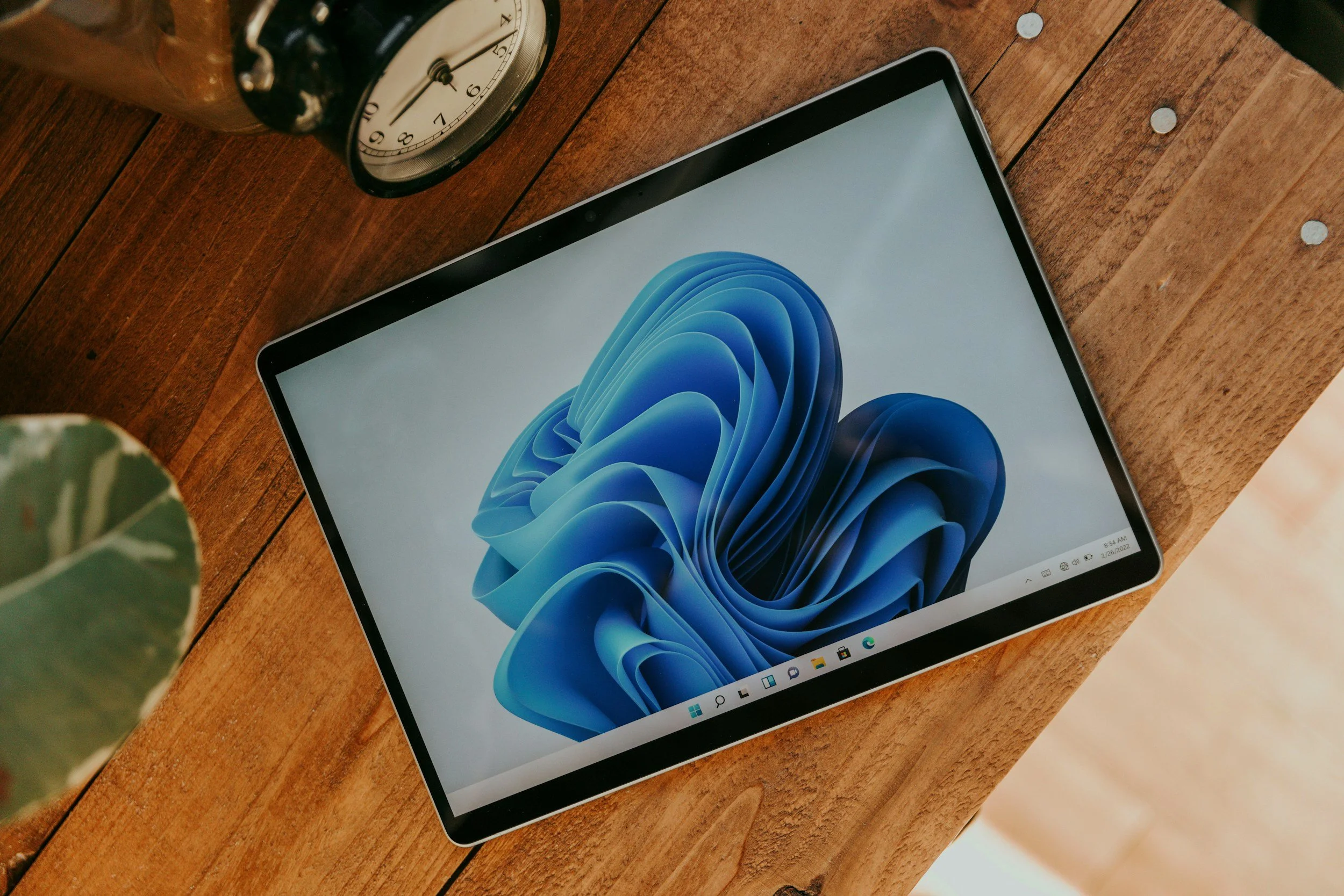 A tablet with the Windows 11 desktop screen displayed, placed on a wooden table. The screen shows a blue swirling abstract pattern.