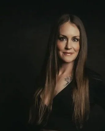 A woman with long brown hair and a tattoo on her chest, wearing a black top, smiling against a dark background.