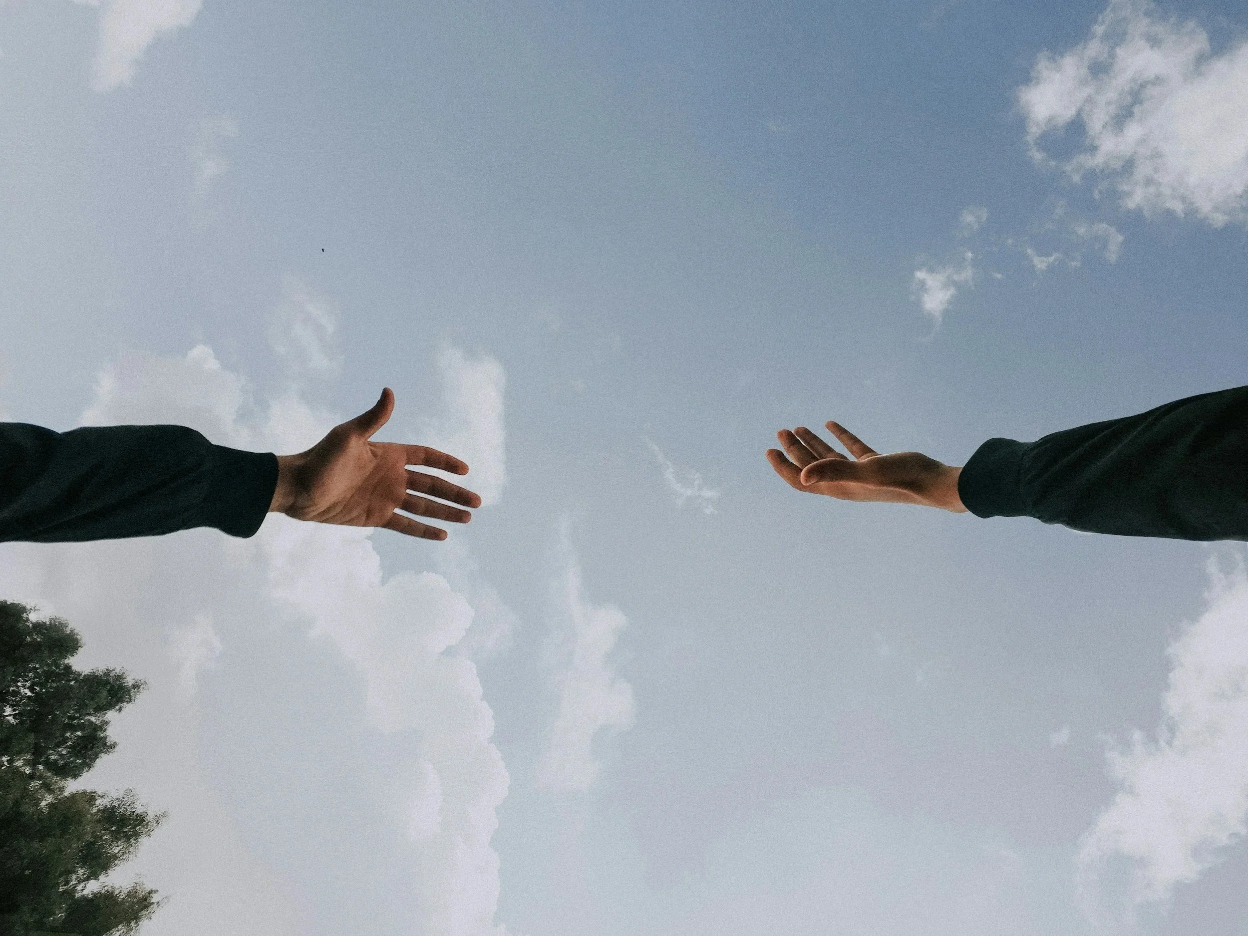Two hands reaching out towards each other in the sky, with clouds and trees visible.