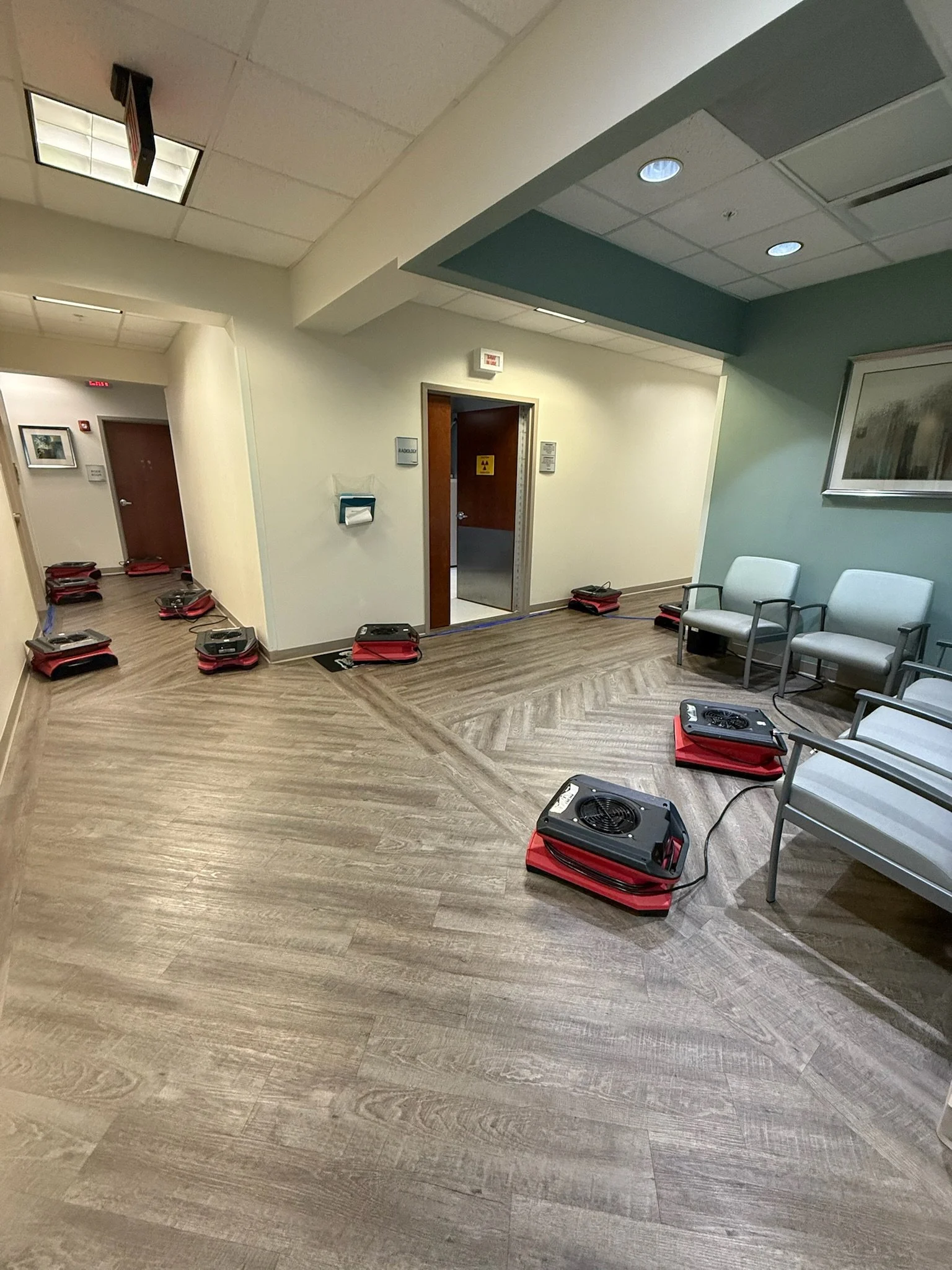 Hospital waiting room with multiple portable air purifiers or fans on the floor, chairs against the wall, and a door with a biohazard sign.