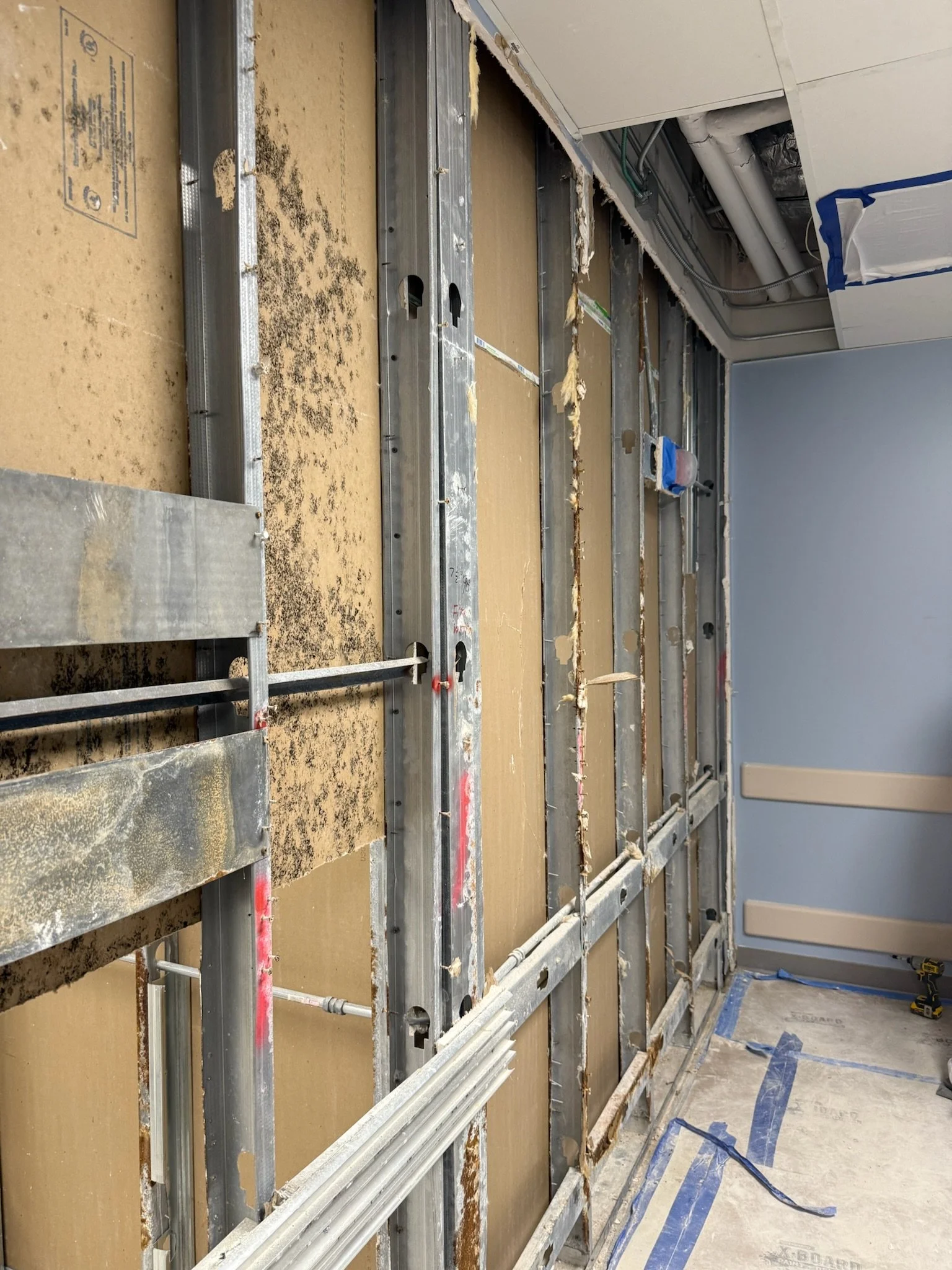 Construction site showing a partially built wall with metal studs, some insulation, and blue painter's tape on the floor, with drywall installation in progress.