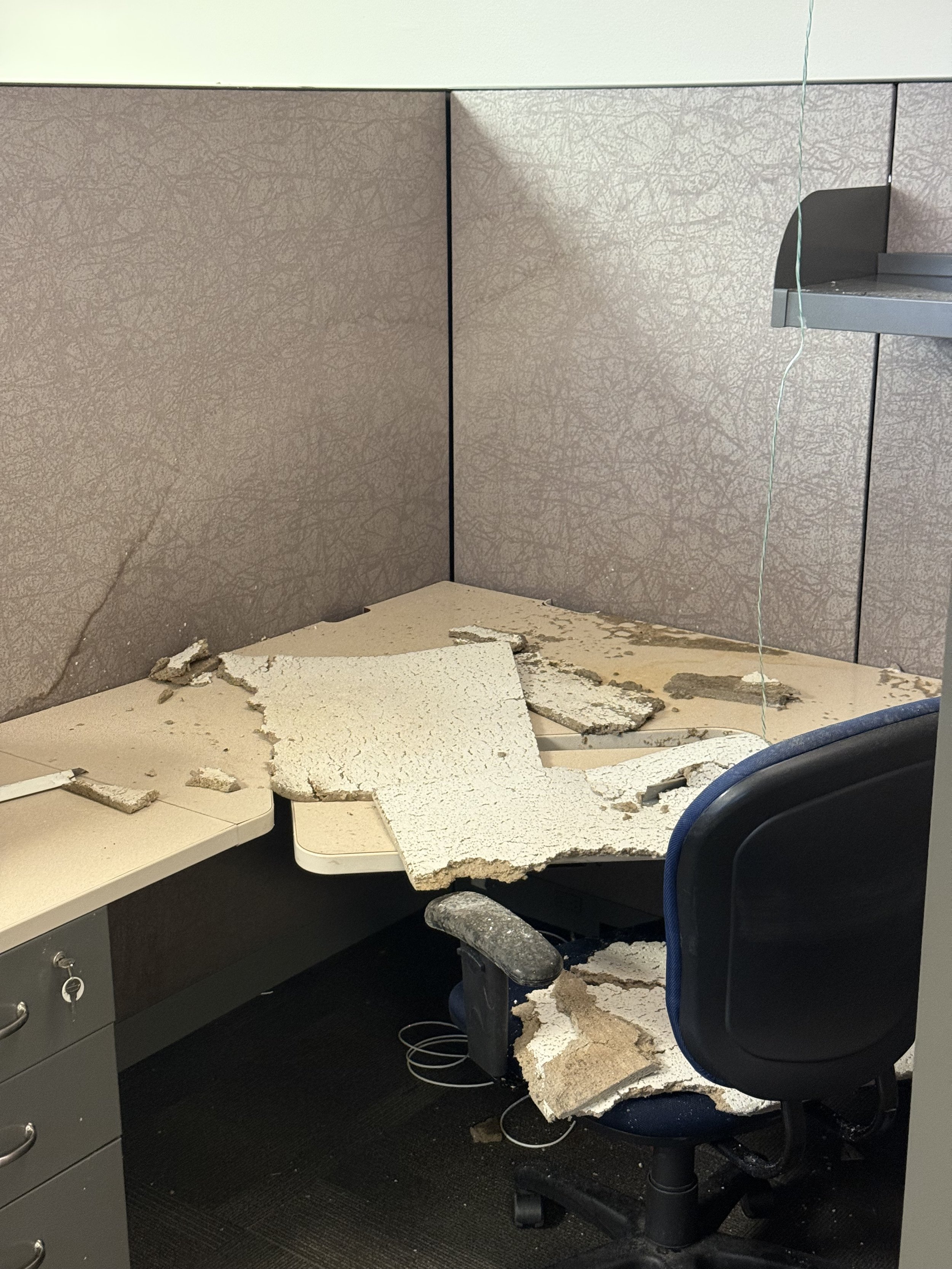 An office cubicle with a damaged desk and broken ceiling tiles on the floor.