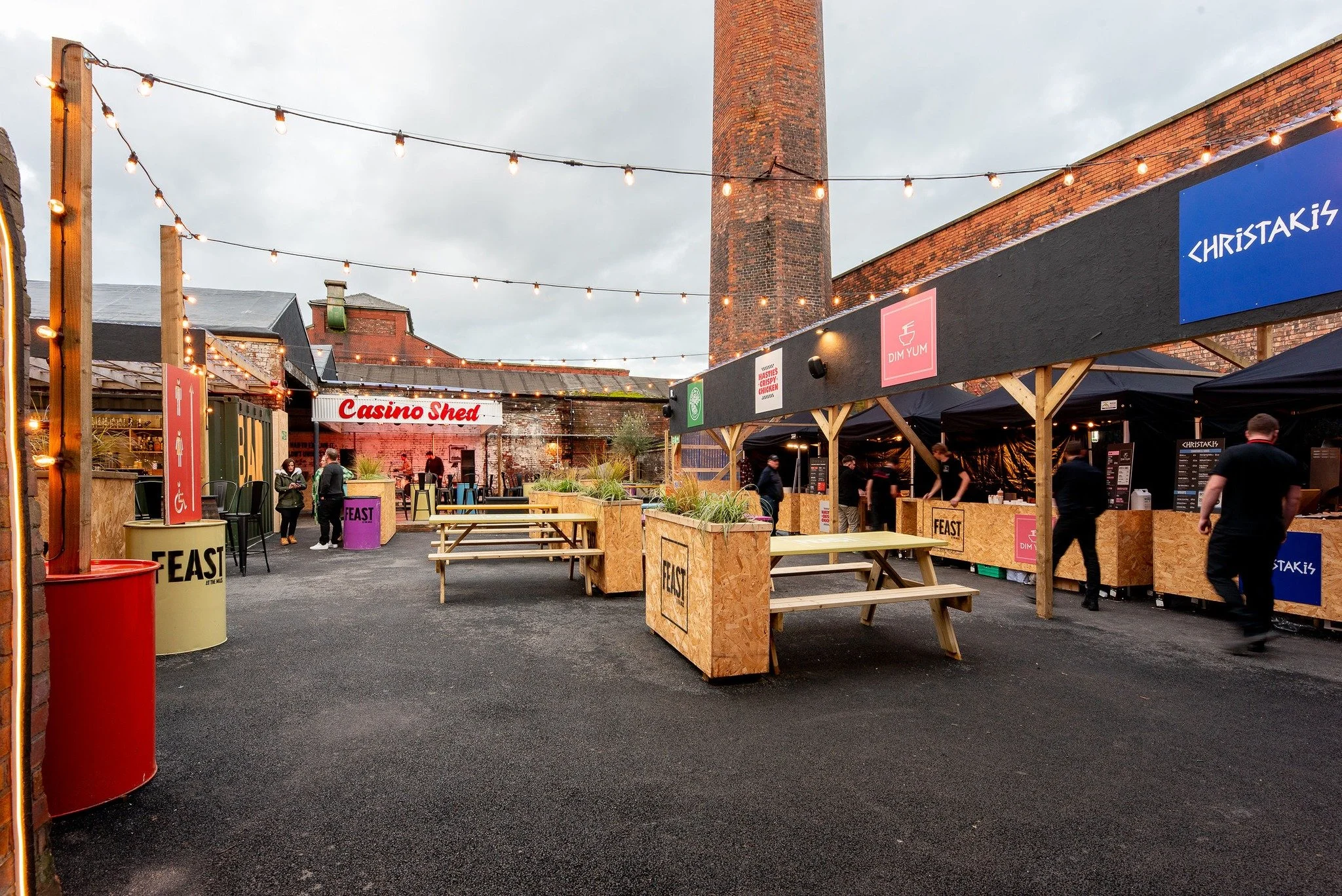 An outdoor food and drink festival with string lights, food stalls, and picnic tables on a paved courtyard. There's a red sign reading "Casino Shed" and various colorful signs on the stalls, Feast at the Mills, Wigan 