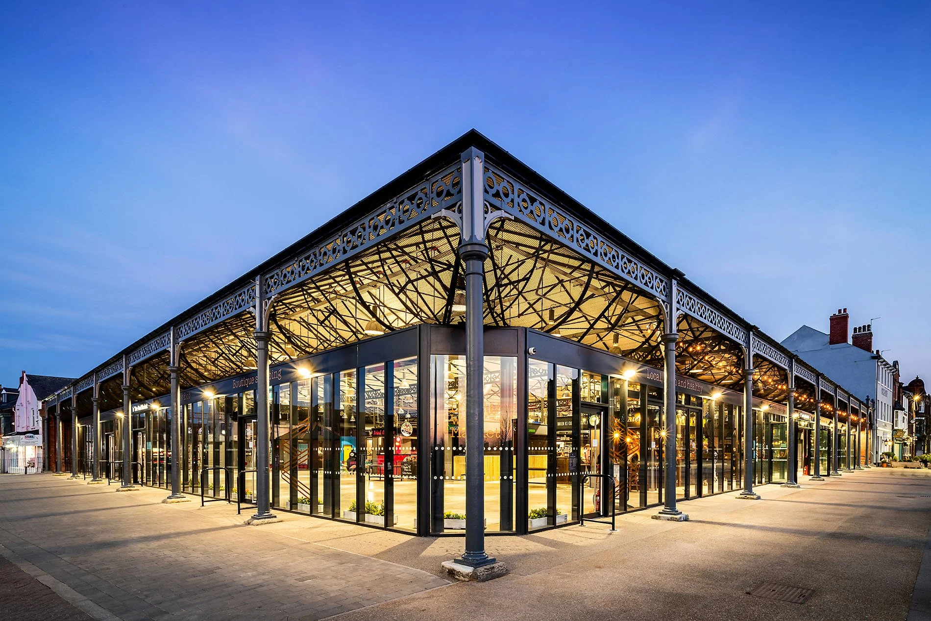 Doncaster Wool Market. A modern shopping mall with glass walls and an ornate metal roof, illuminated inside during dusk, located in a small town.