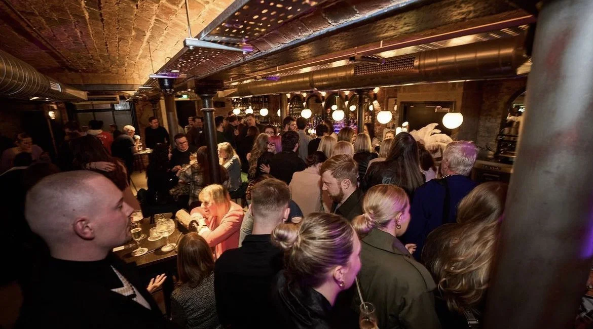Crowded bar or restaurant with people socializing, warm lighting, exposed brick walls, and industrial decor.