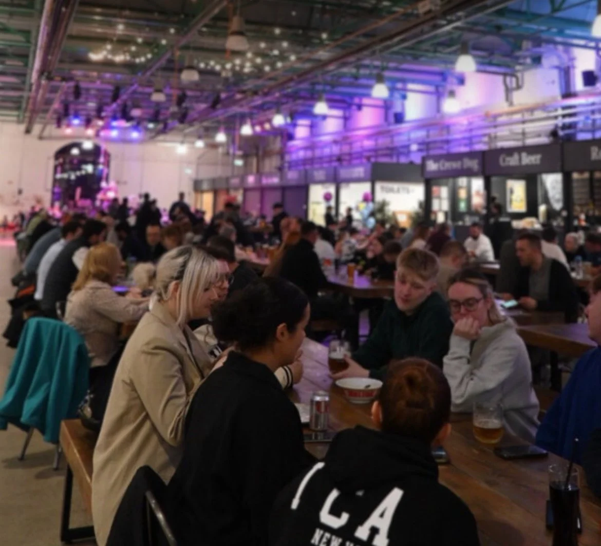 Crewe Market Hall. People sitting at tables in a large indoor beer hall or food court with colorful lighting and food vendors in the background.