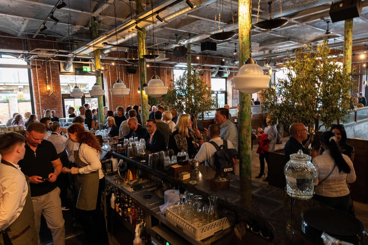 A busy indoor bar and restaurant with numerous patrons socializing and dining. The space features exposed brick walls, large windows, and modern lighting fixtures, with green-painted decorative pillars and some potted plants.
