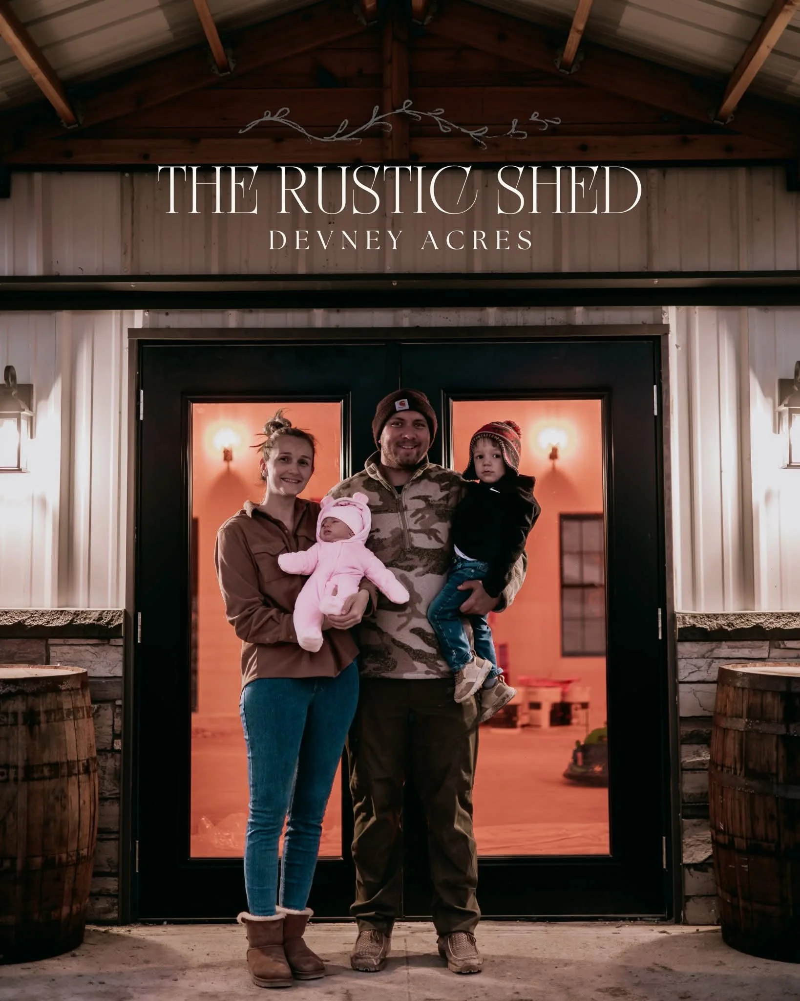 A family of four standing in front of a rustic shed with a sign that reads 'The Rustic Shed, Devney Acres.' The family is smiling; the mother is holding a baby in pink, the father is holding a young boy, and they are dressed warmly.