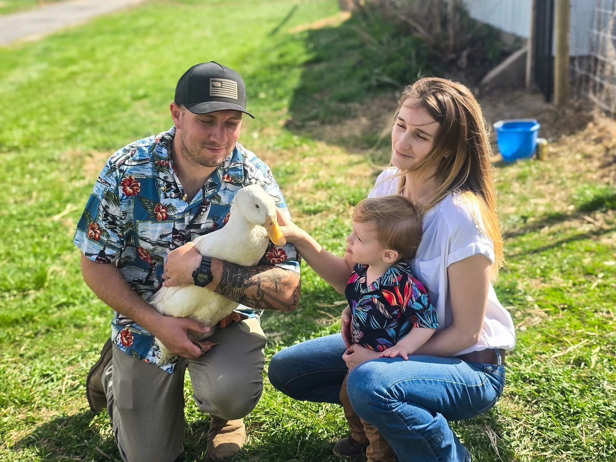 Family outdoors, petting a white duck, with a man in a Hawaiian shirt, a woman and a young girl, in a grassy backyard.