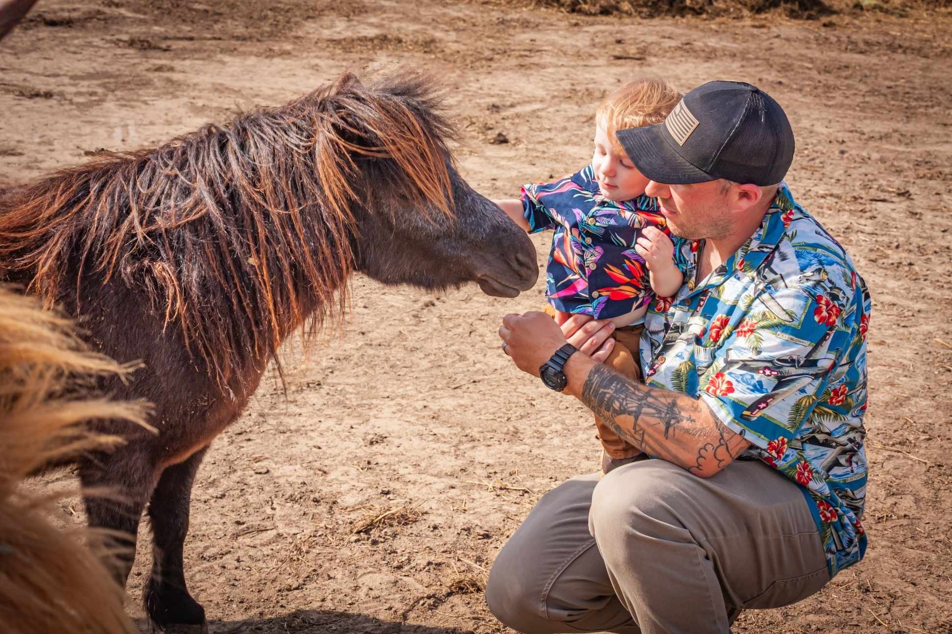 A man with tattoos on his arm and wearing a floral Hawaiian shirt kneels on dirt ground holding a young boy in his arms, both petting a brown pinto horse with a long mane.