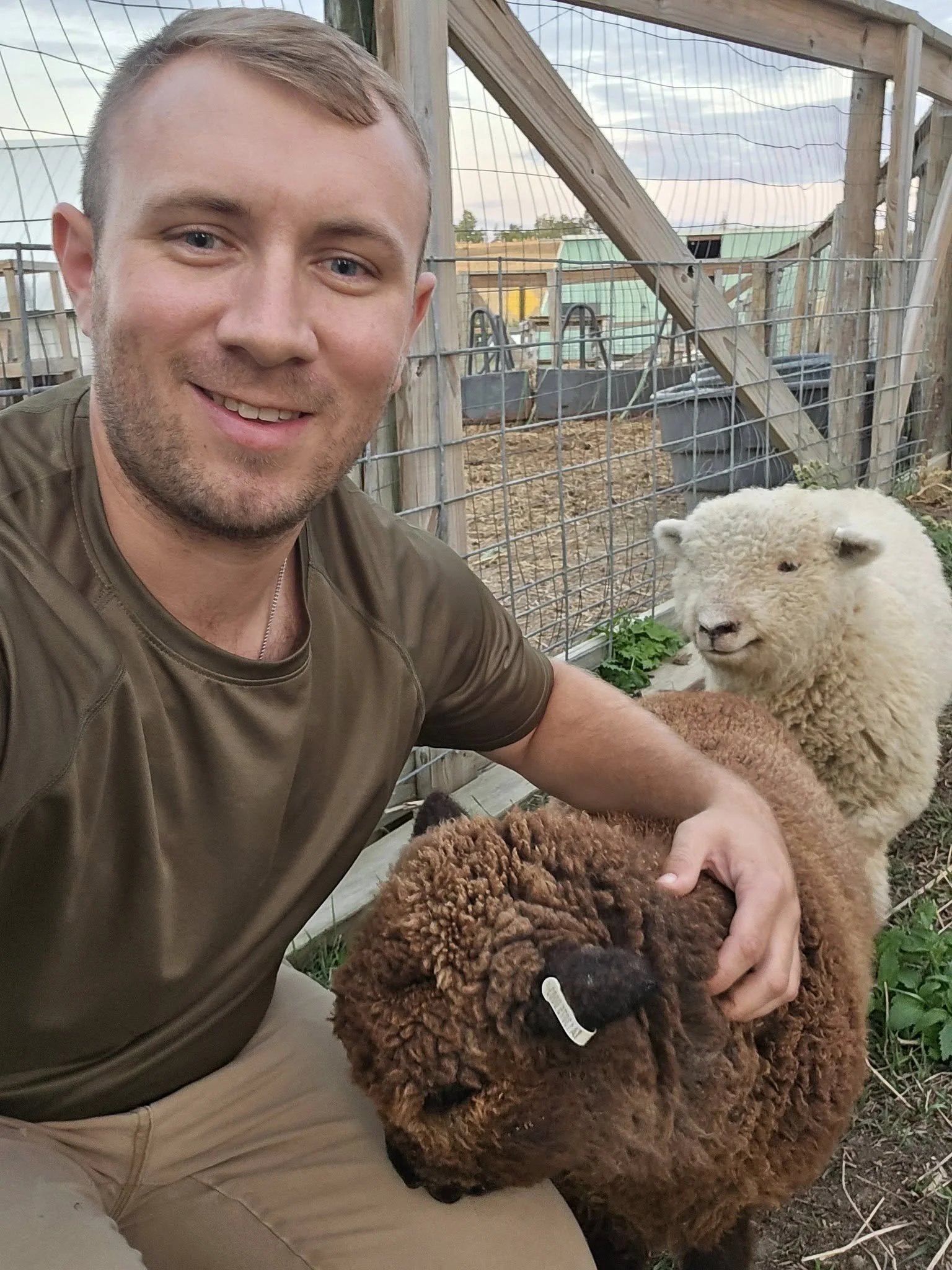 A smiling man in an outdoor farm setting with a brown sheep on his lap and a white sheep behind him.