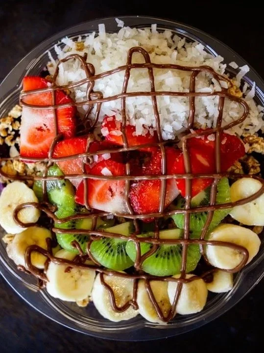 A clear plastic bowl filled with sliced bananas, kiwi, strawberries, and other fruits, topped with shredded coconut and drizzled with chocolate sauce on a dark wooden surface.