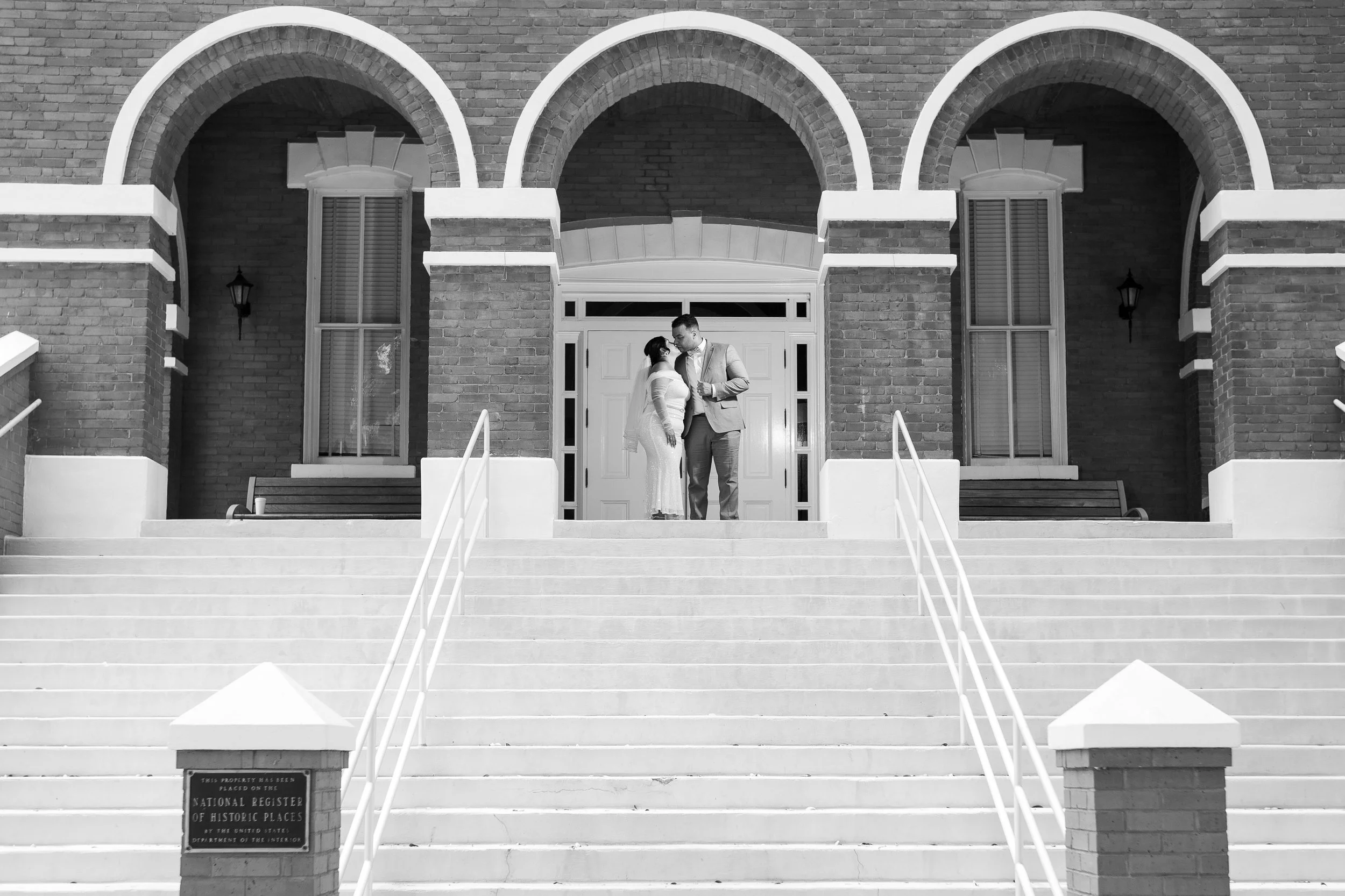 A couple with the woman in a wedding dress and the man in a suit standing on the steps of a historic brick building, sharing a moment together.