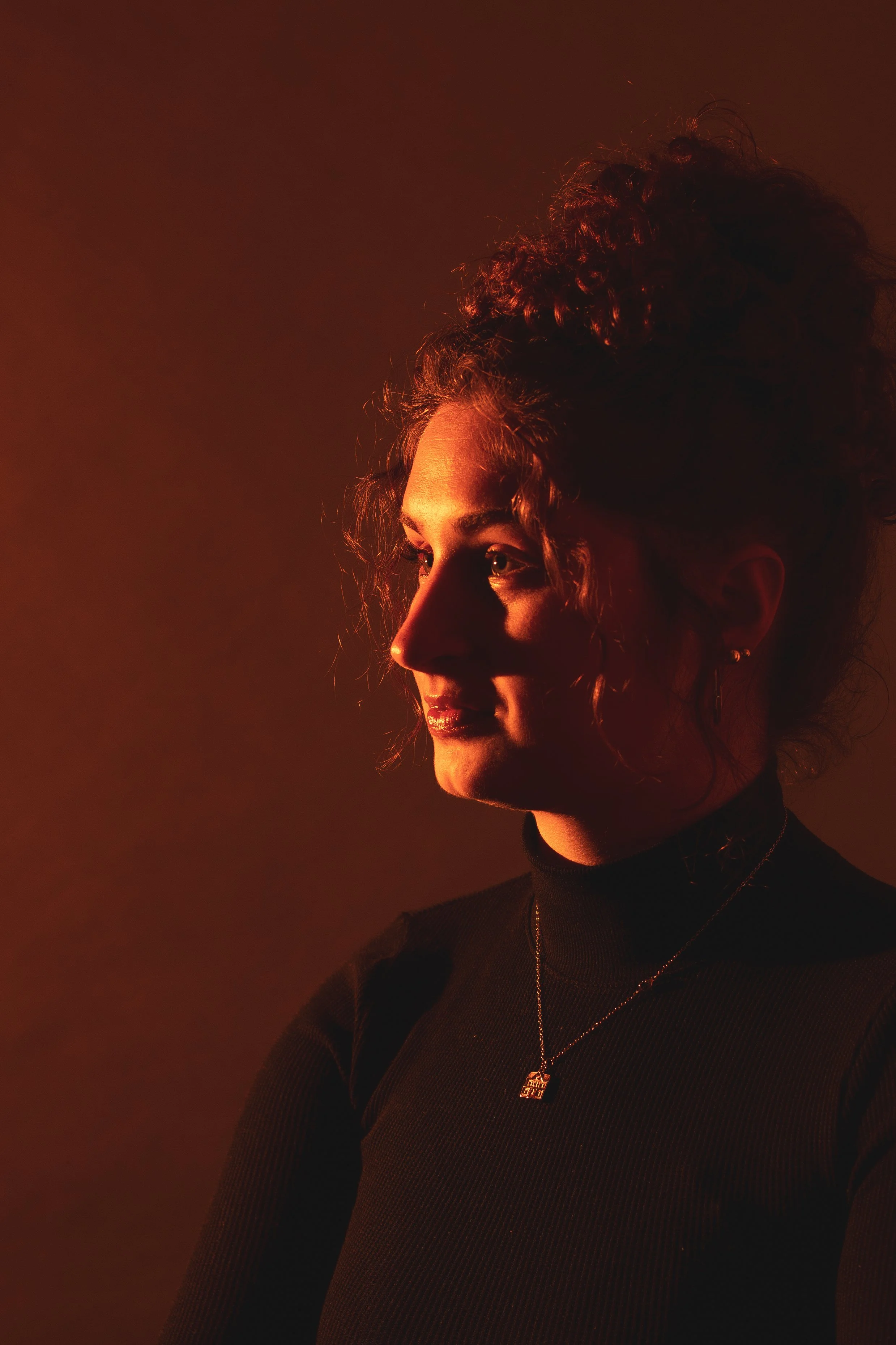 Side profile of a woman with curly hair in a bun, wearing a black turtleneck and a necklace, illuminated by warm, soft lighting.