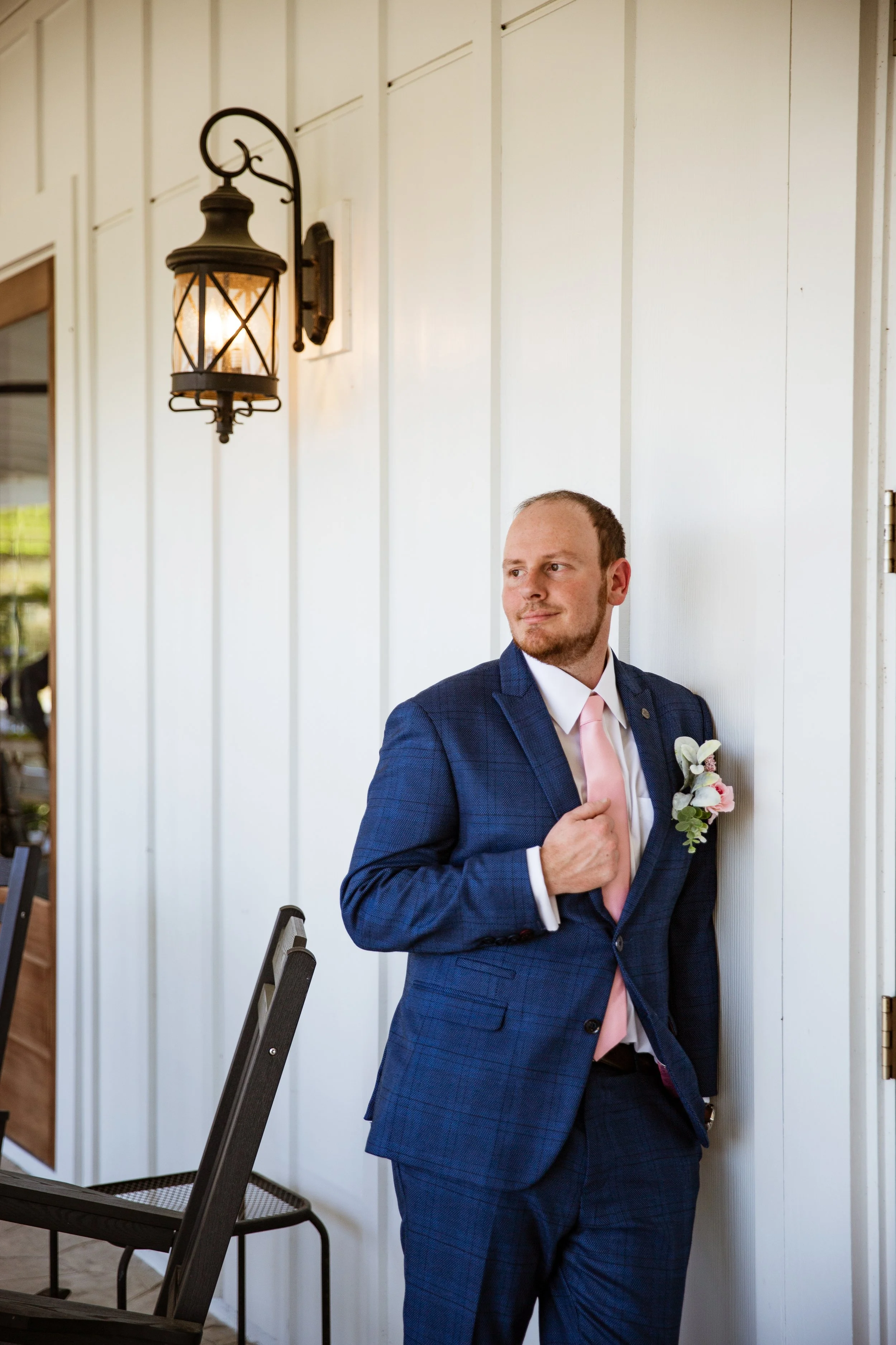 Man in a blue checkered suit with a pink tie and flower boutonniere leaning against a white wall next to an outdoor light fixture.