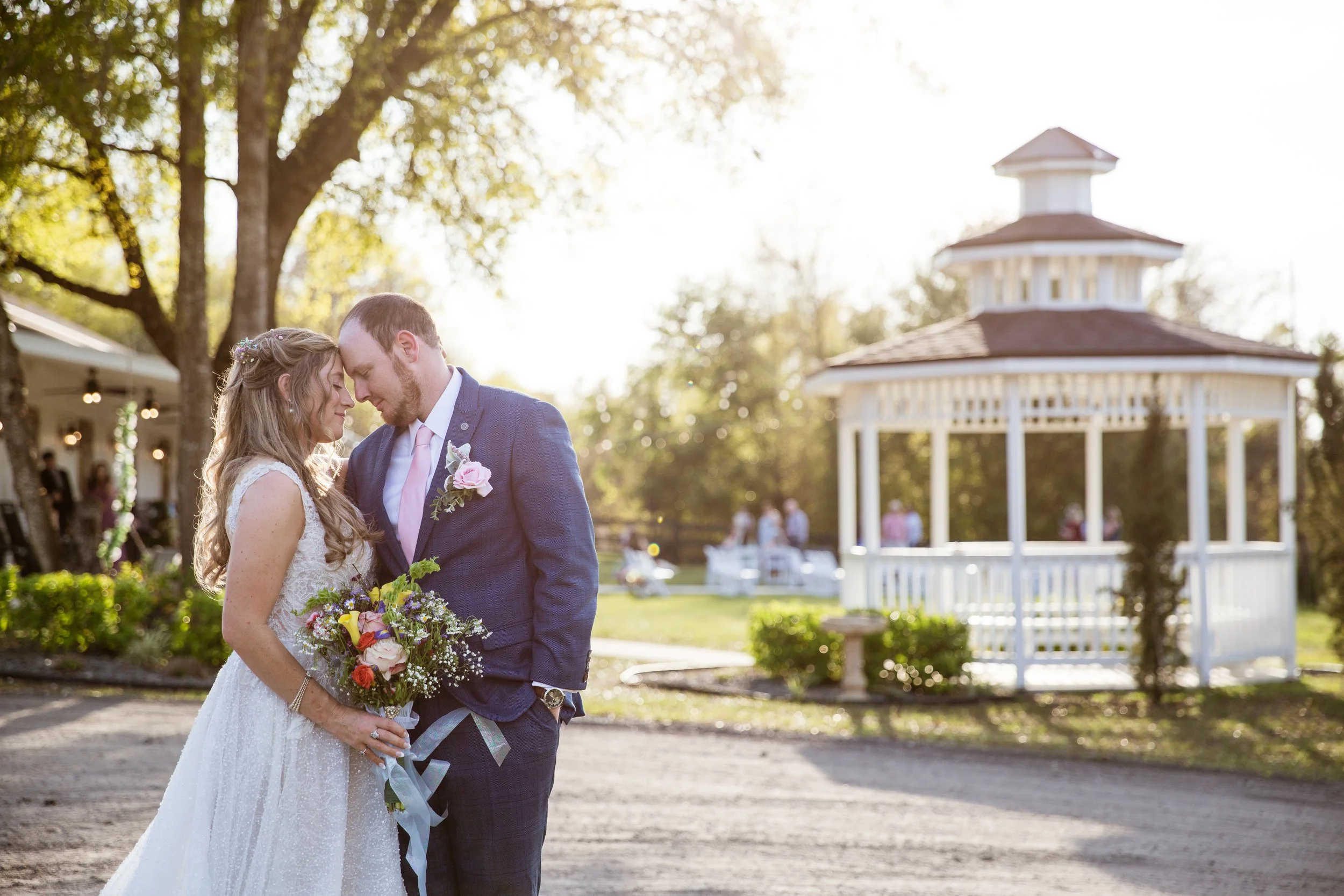 A bride and groom sharing a close moment outdoors in the evening sun, with greenery and a white gazebo in the background.