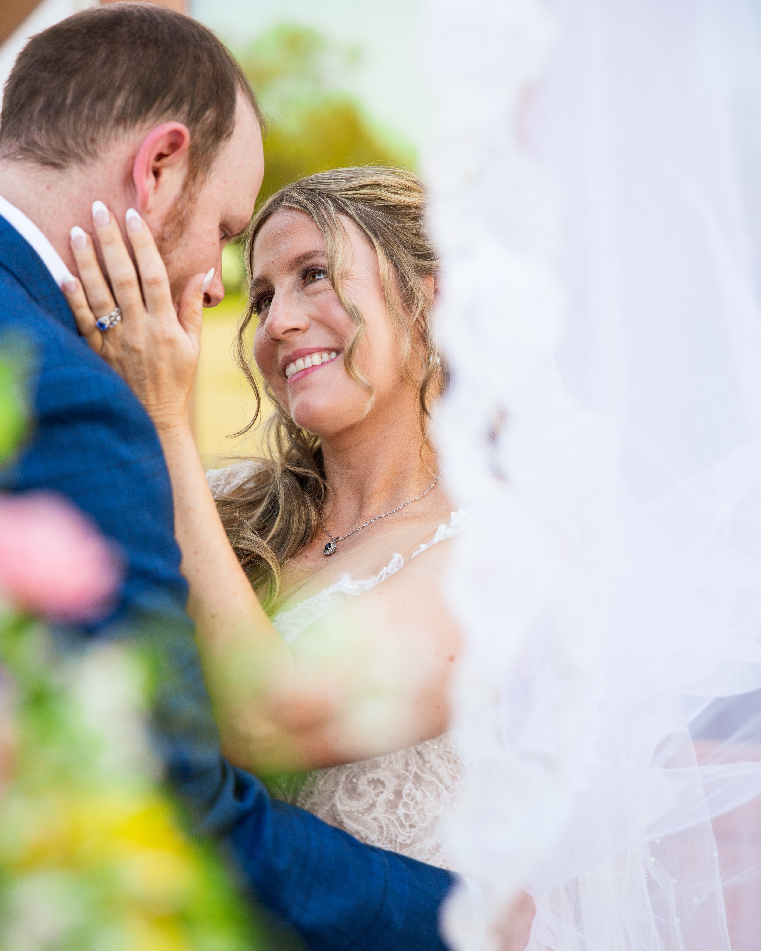 A bride and groom gazing at each other, with the bride smiling and the groom holding her face, surrounded by flowers.