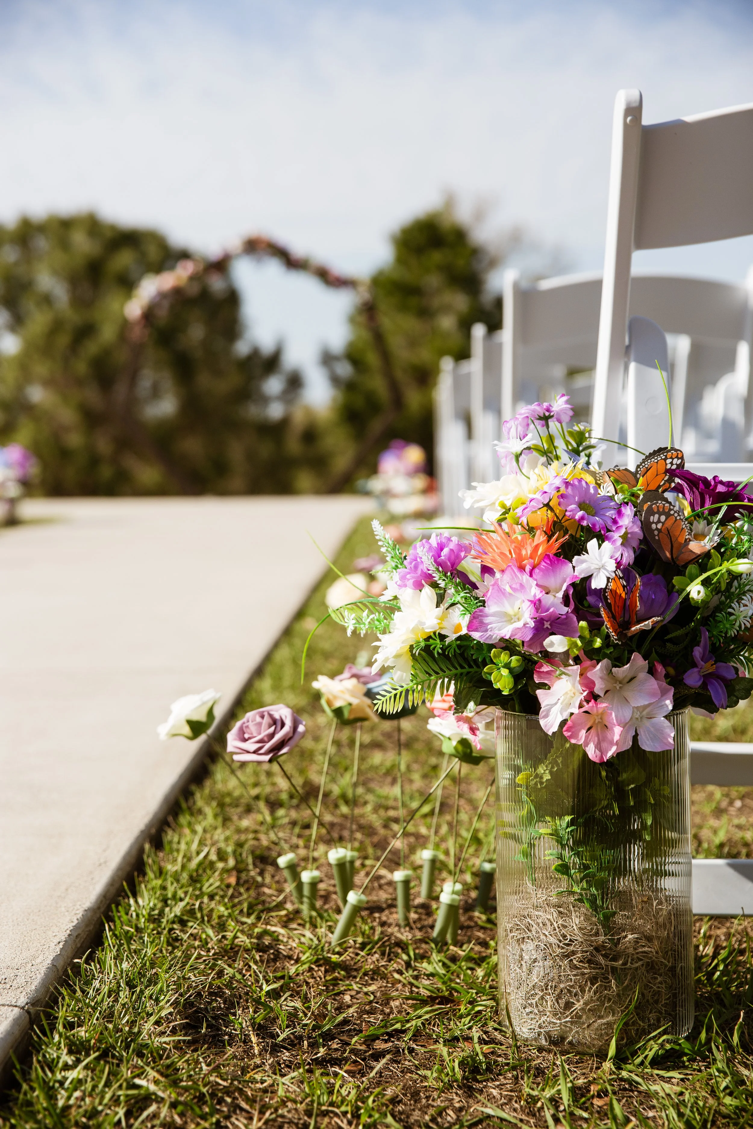A flower arrangement in a clear vase at a wedding ceremony setup with white chairs and an arch in the background.