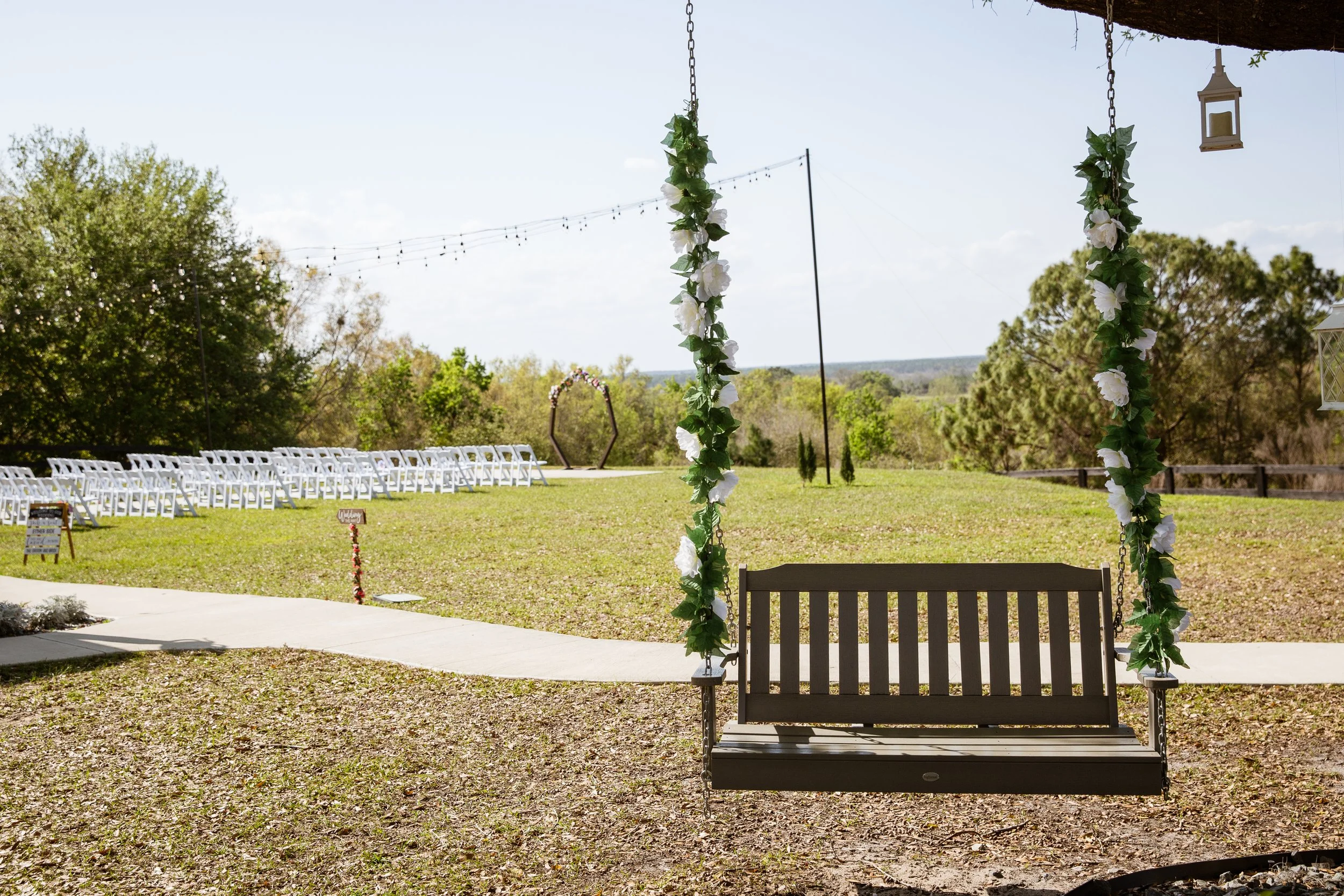 An outdoor wedding setup with white chairs arranged in rows on a grassy area, a decorated swing with flowers in the foreground, and a pathway leading to a wooden arch in the distance under a clear sky.
