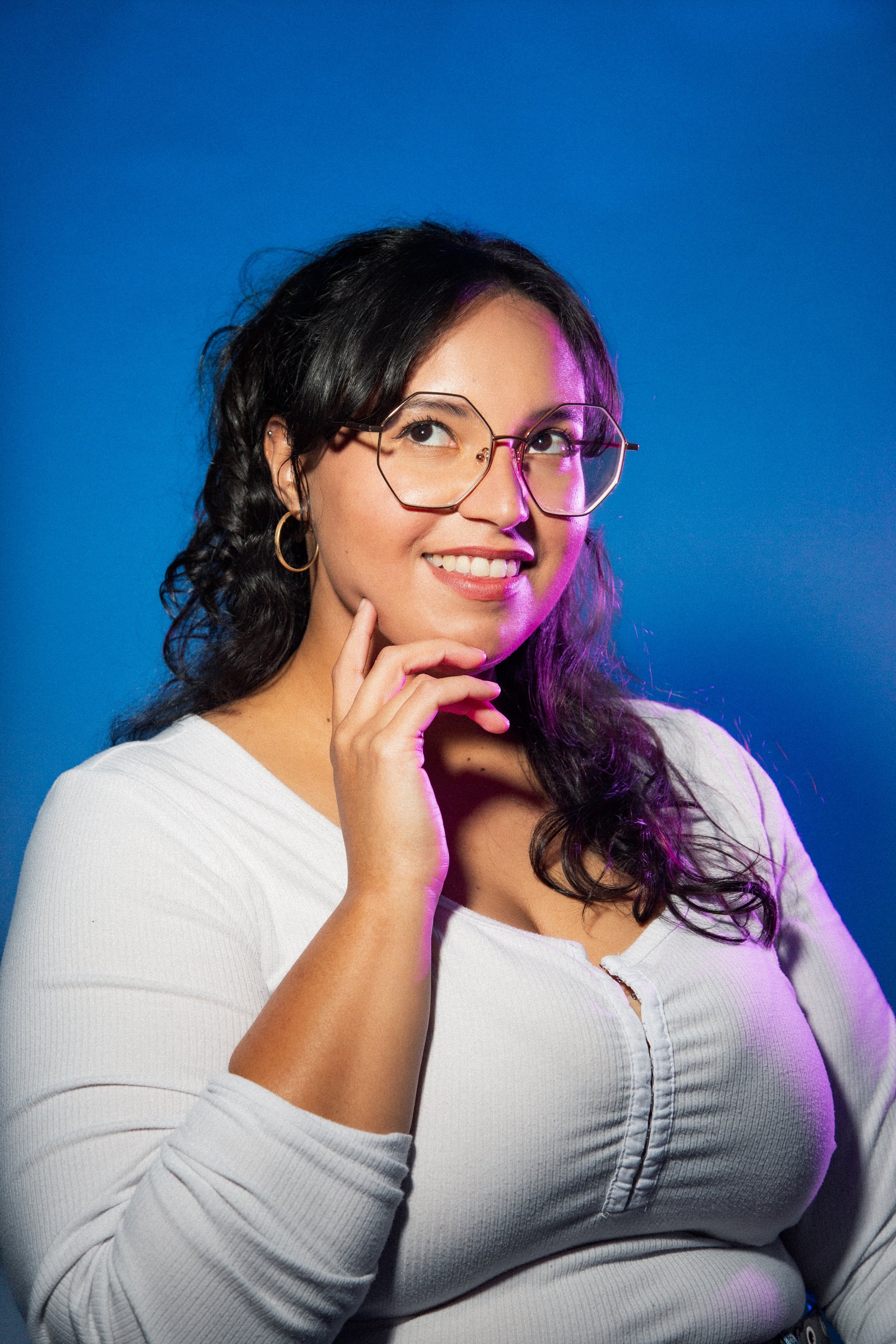 A woman with dark hair wearing glasses, gold hoop earrings, and a white top, smiling thoughtfully against a blue background illuminated with purple lighting.