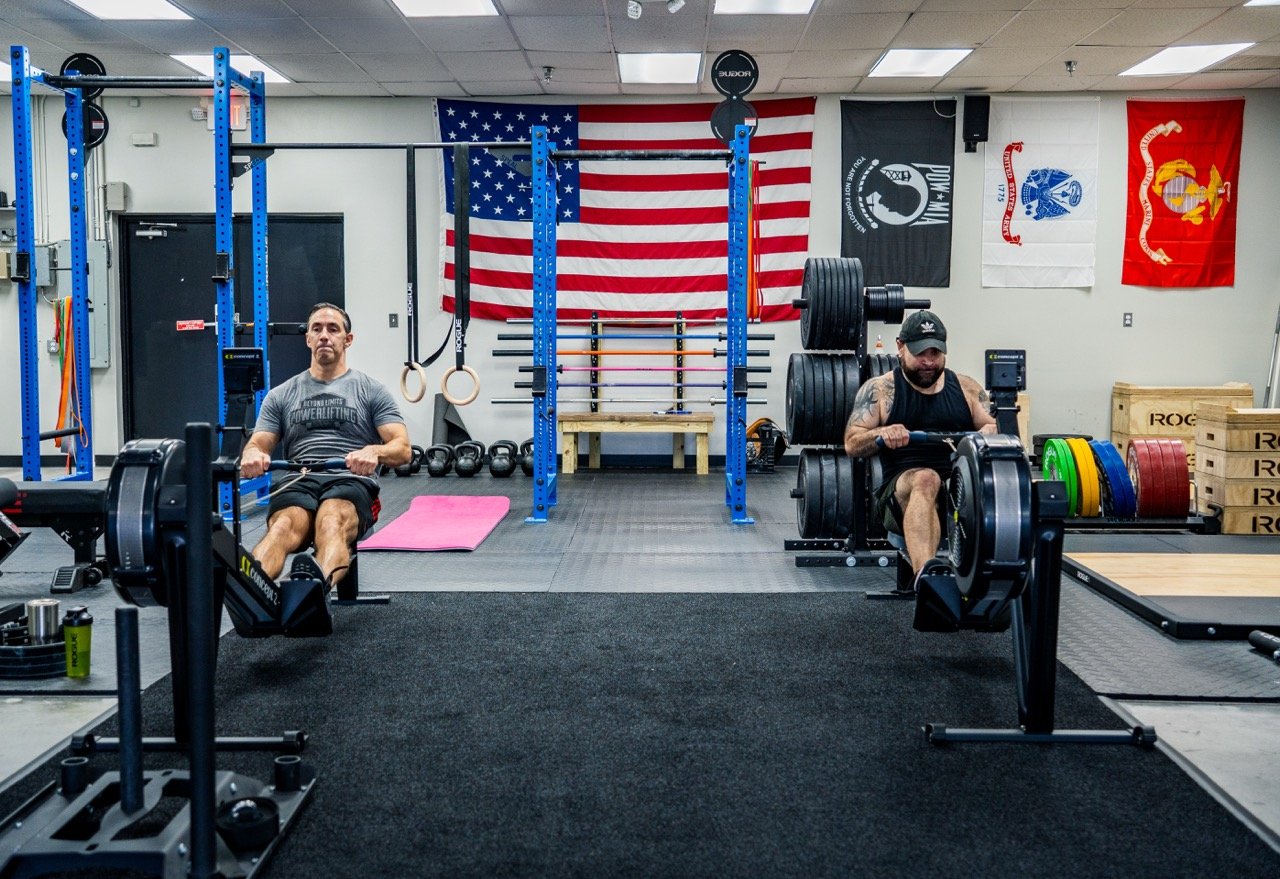 Two men working out with rowing machines in a gym with American flags and others on the wall.