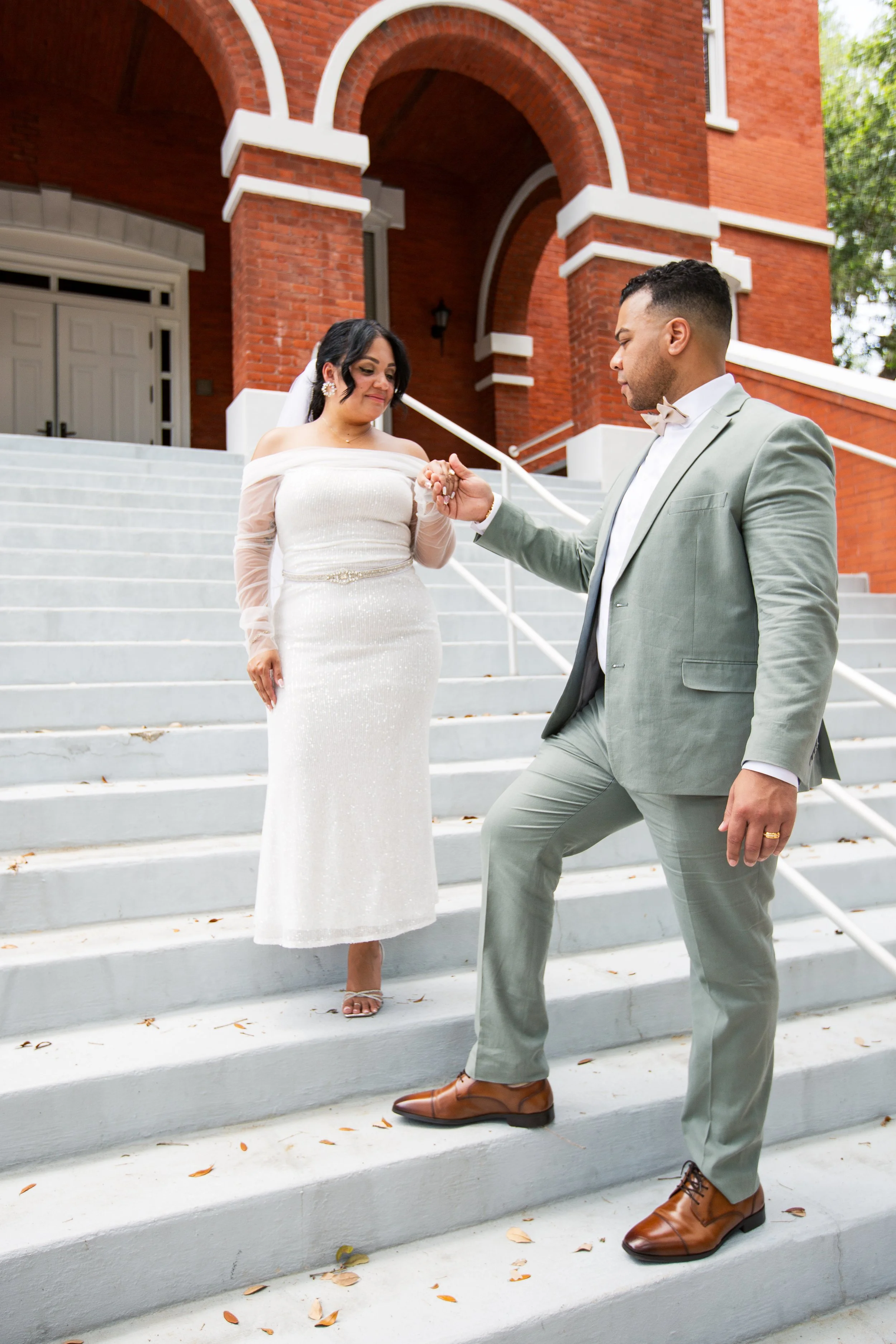 A groom in a light green suit kneeling and holding the hand of a bride in a white wedding dress on outdoor steps of a red brick building.