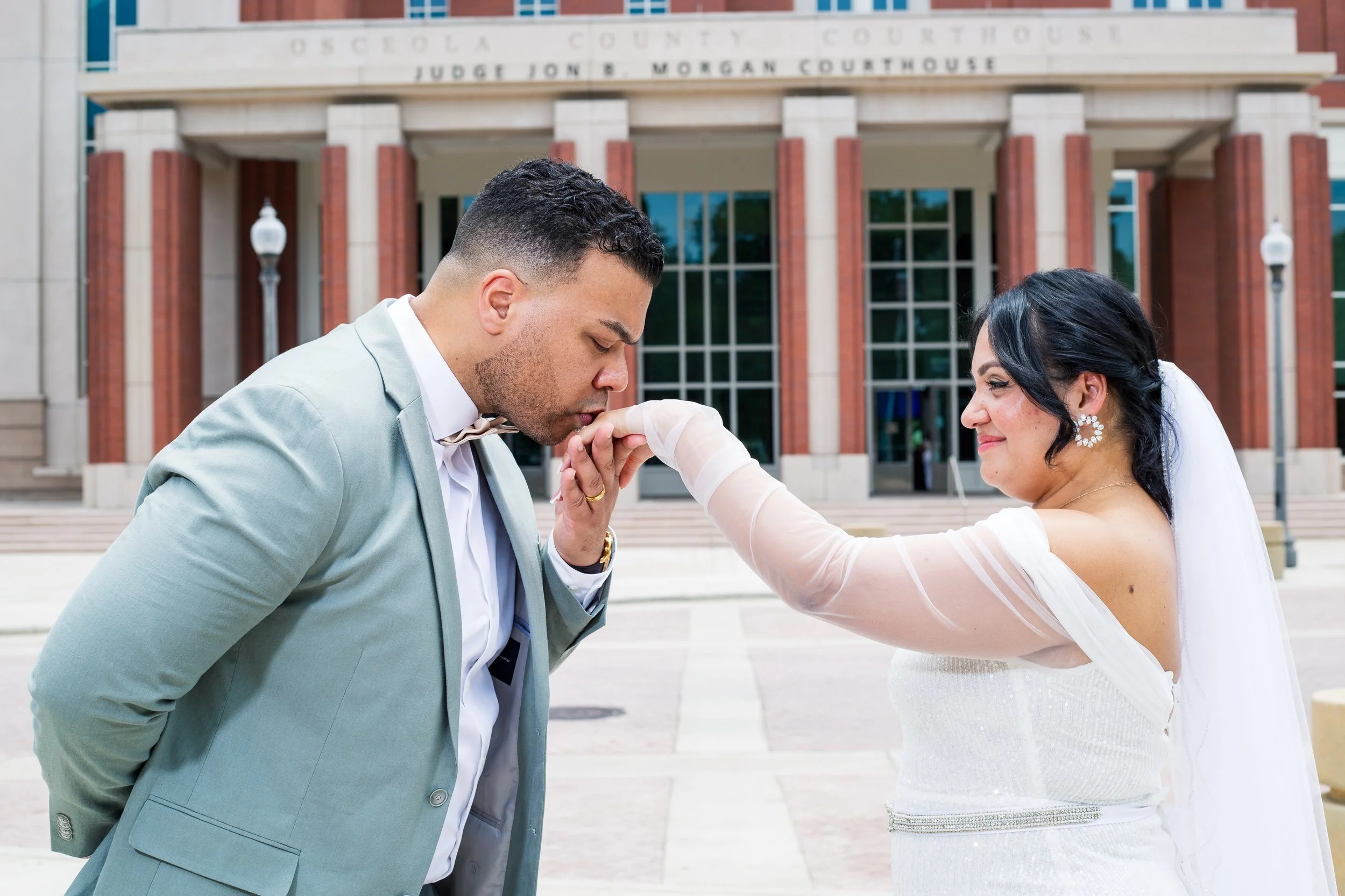 A woman in a white wedding dress is kissing a man in a light gray suit on the hand in front of a courthouse building.