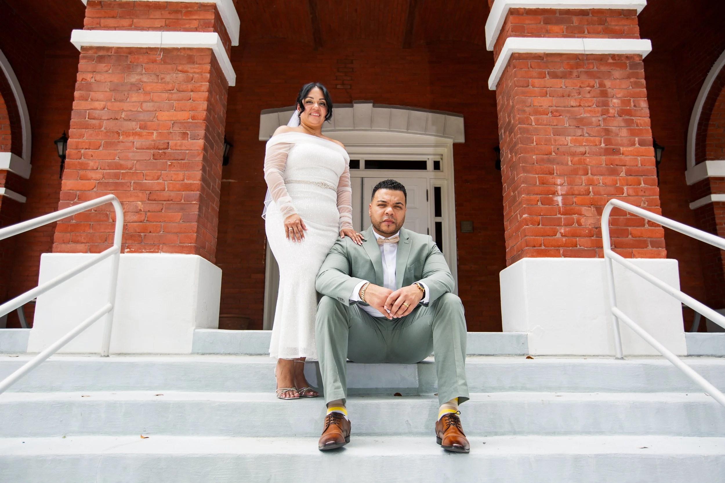 A woman in a white wedding dress and a man in a light green suit with a bowtie sitting and standing on the steps of a red brick building.