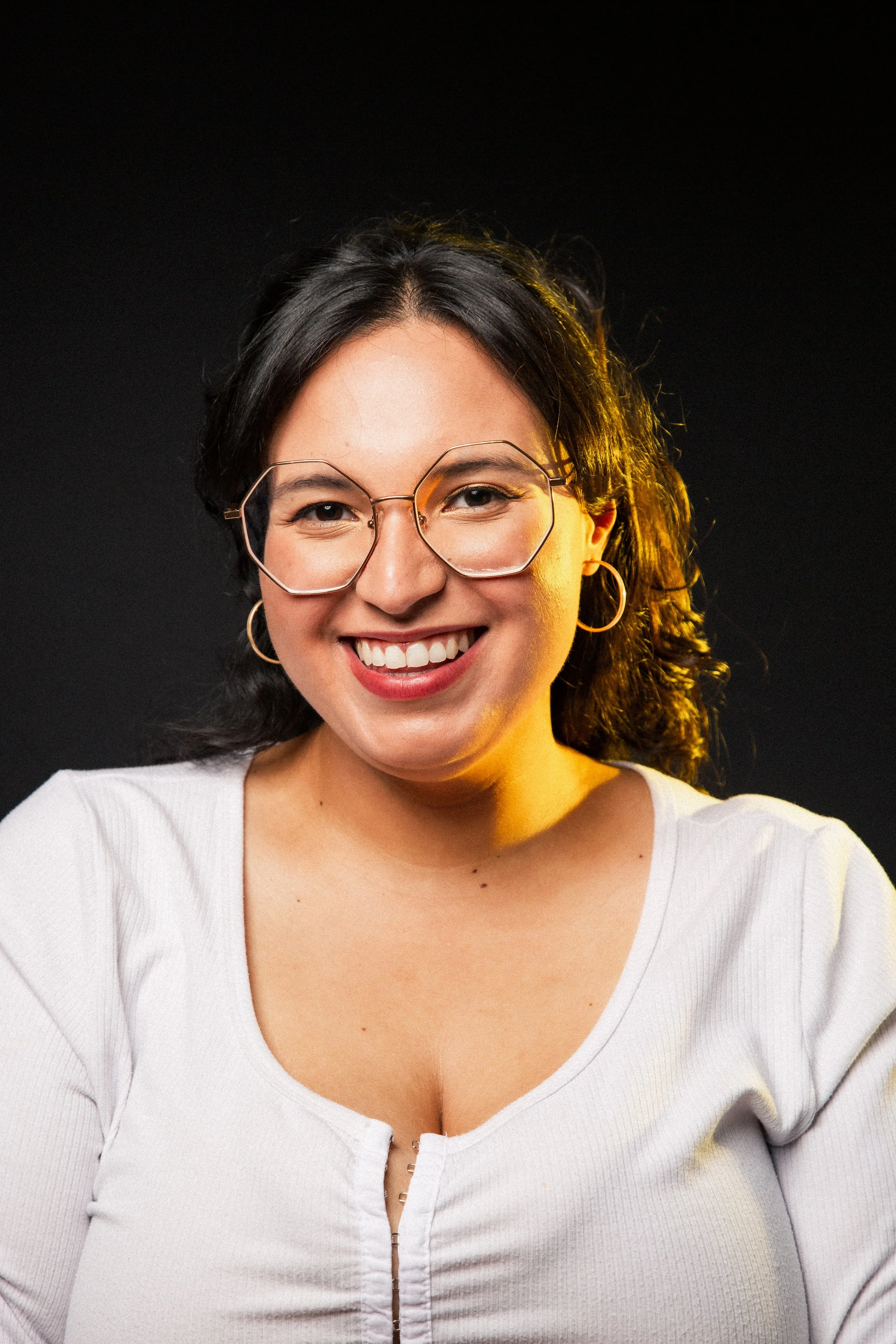 A smiling woman with black hair, wearing large hexagonal glasses, hoop earrings, and a white top, standing against a dark background with warm lighting.