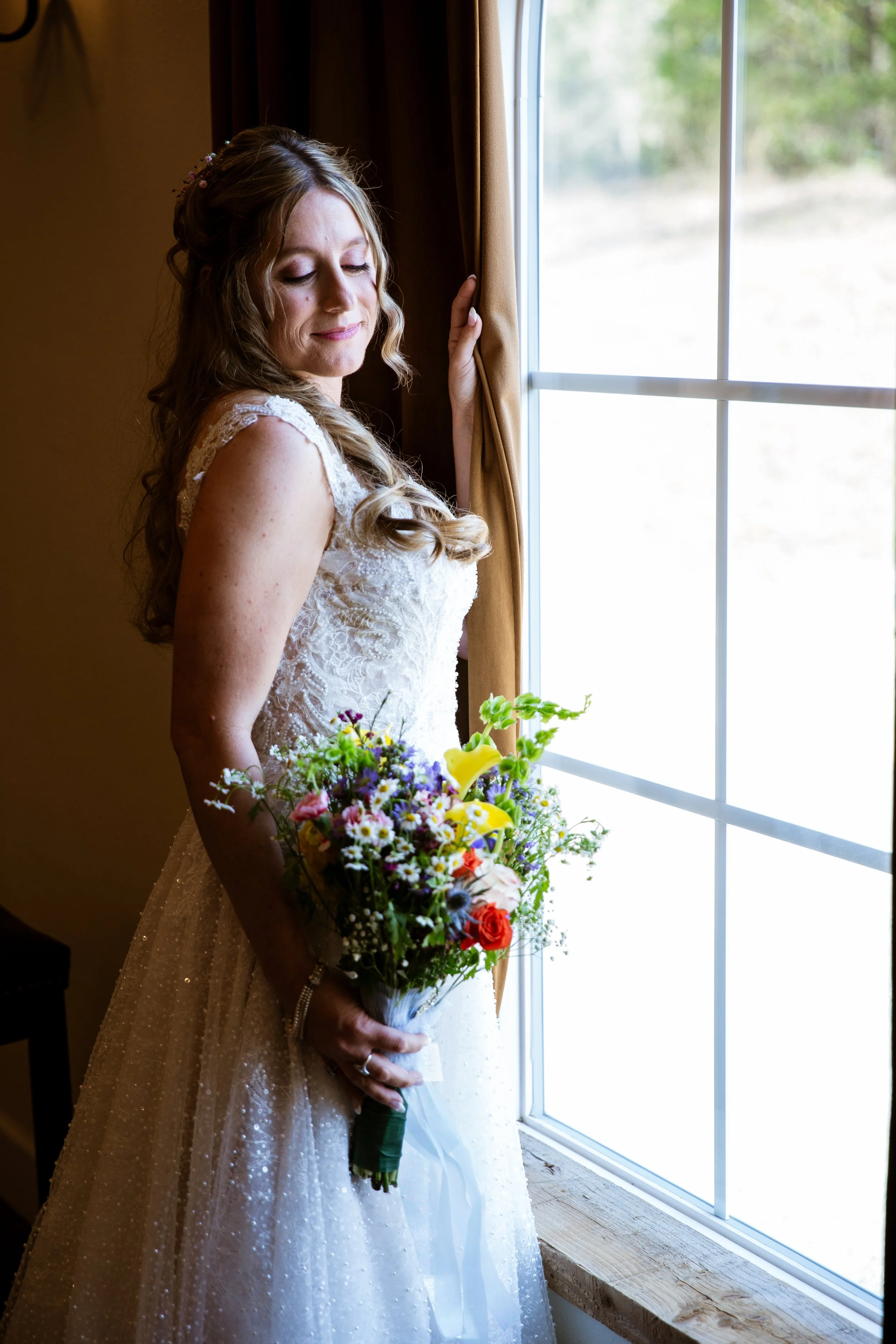 A bride standing by a window holding a bouquet of colorful flowers, wearing a lace wedding dress and smiling softly.