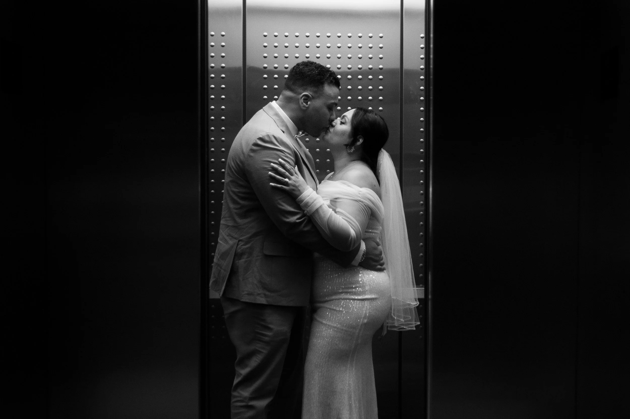 A black and white photo of a couple kissing in an elevator. The man is wearing a suit, and the woman is wearing a wedding dress and veil.