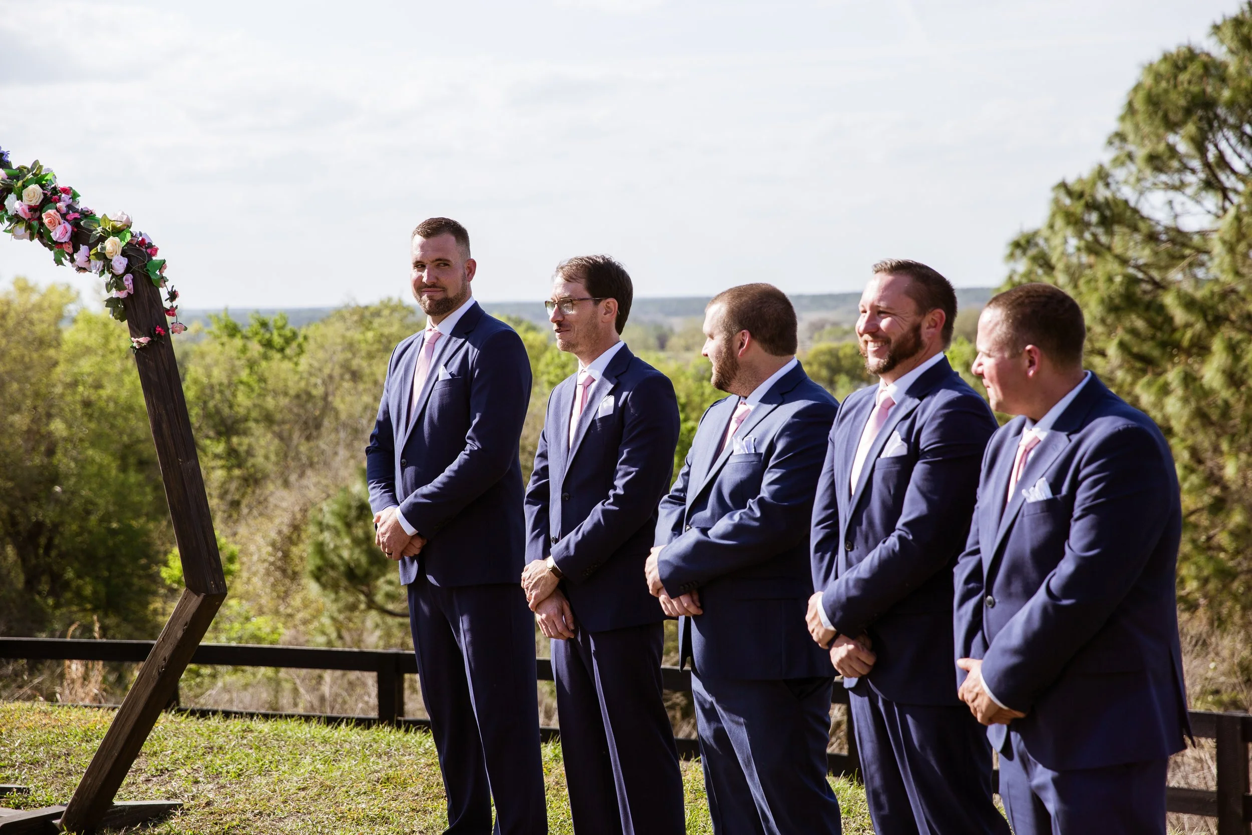 Group of five men in navy suits at an outdoor wedding ceremony with a wooden arch decorated with pink and white flowers, green trees, and open sky in the background.
