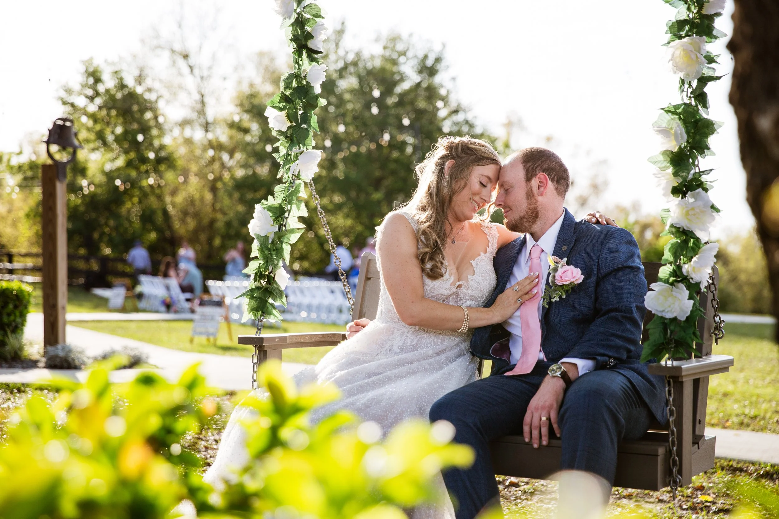 A bride and groom sit on a decorated swing outdoors, sharing a loving moment, with wedding guests in the background.
