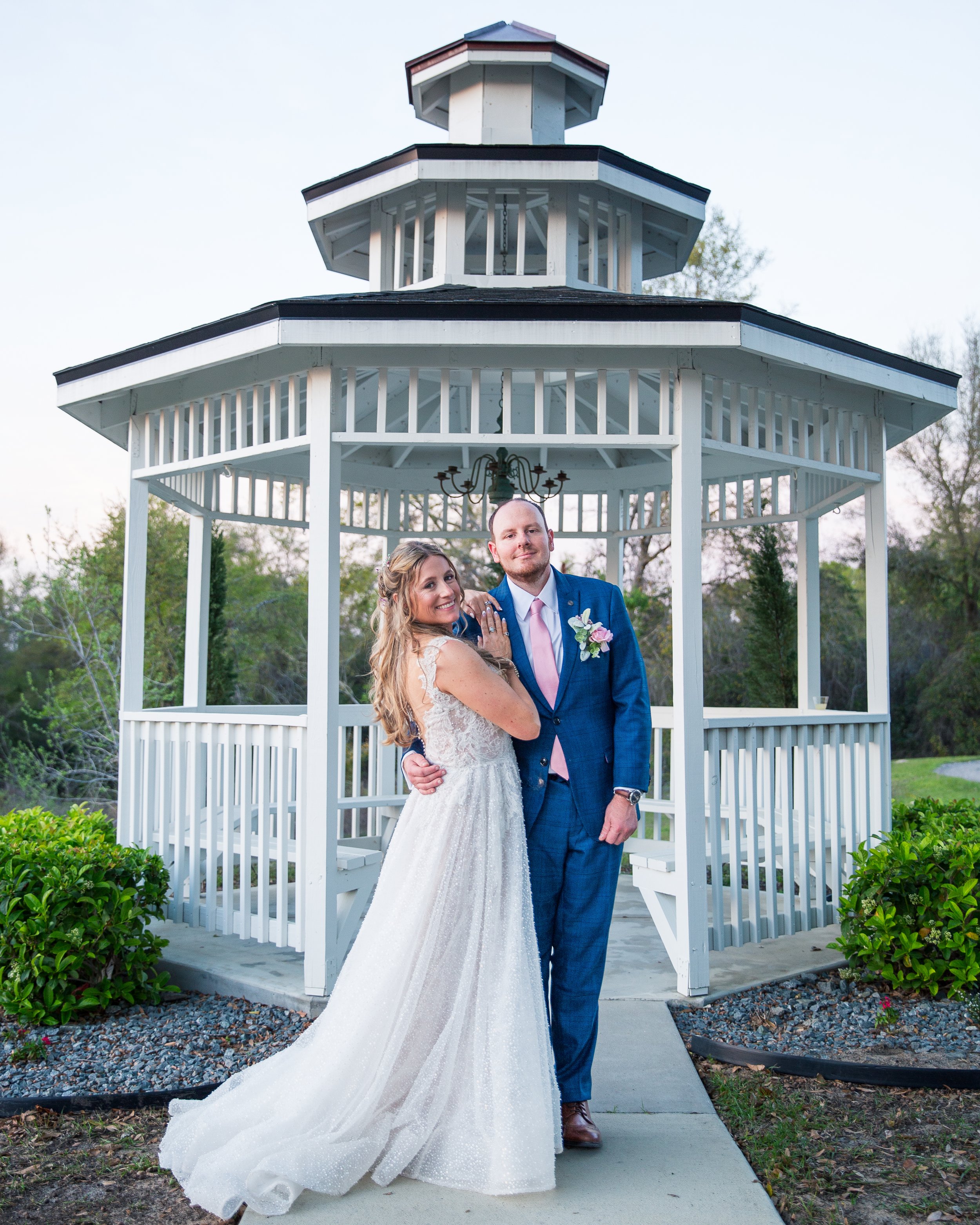 A newlywed couple in wedding attire standing under a white gazebo outdoors, with greenery in the background.