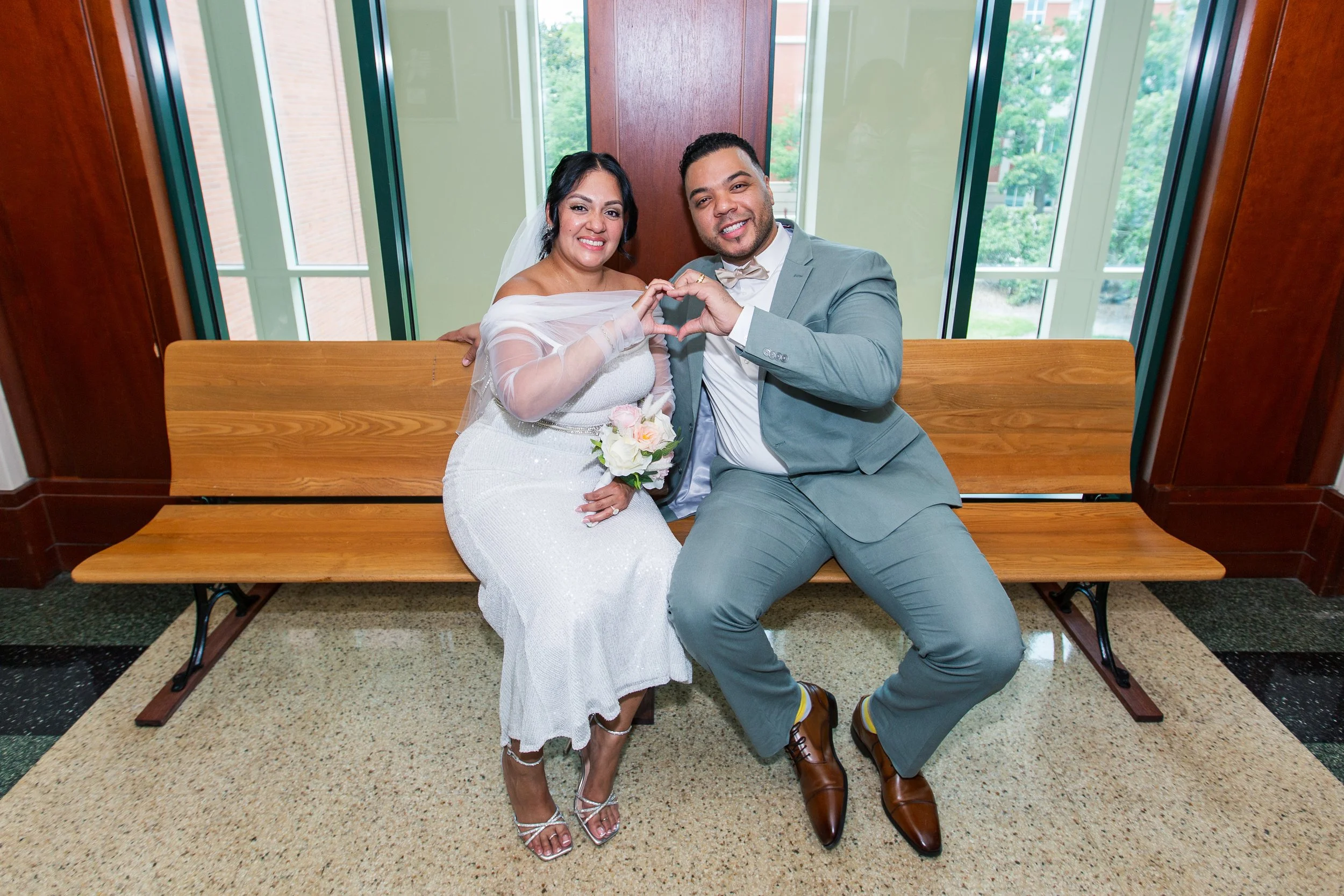A newlywed couple sitting on a wooden bench inside a building, making a heart shape with their hands. The bride is wearing a white wedding dress and holding a bouquet, while the groom is dressed in a grey suit with brown shoes. They are smiling and s