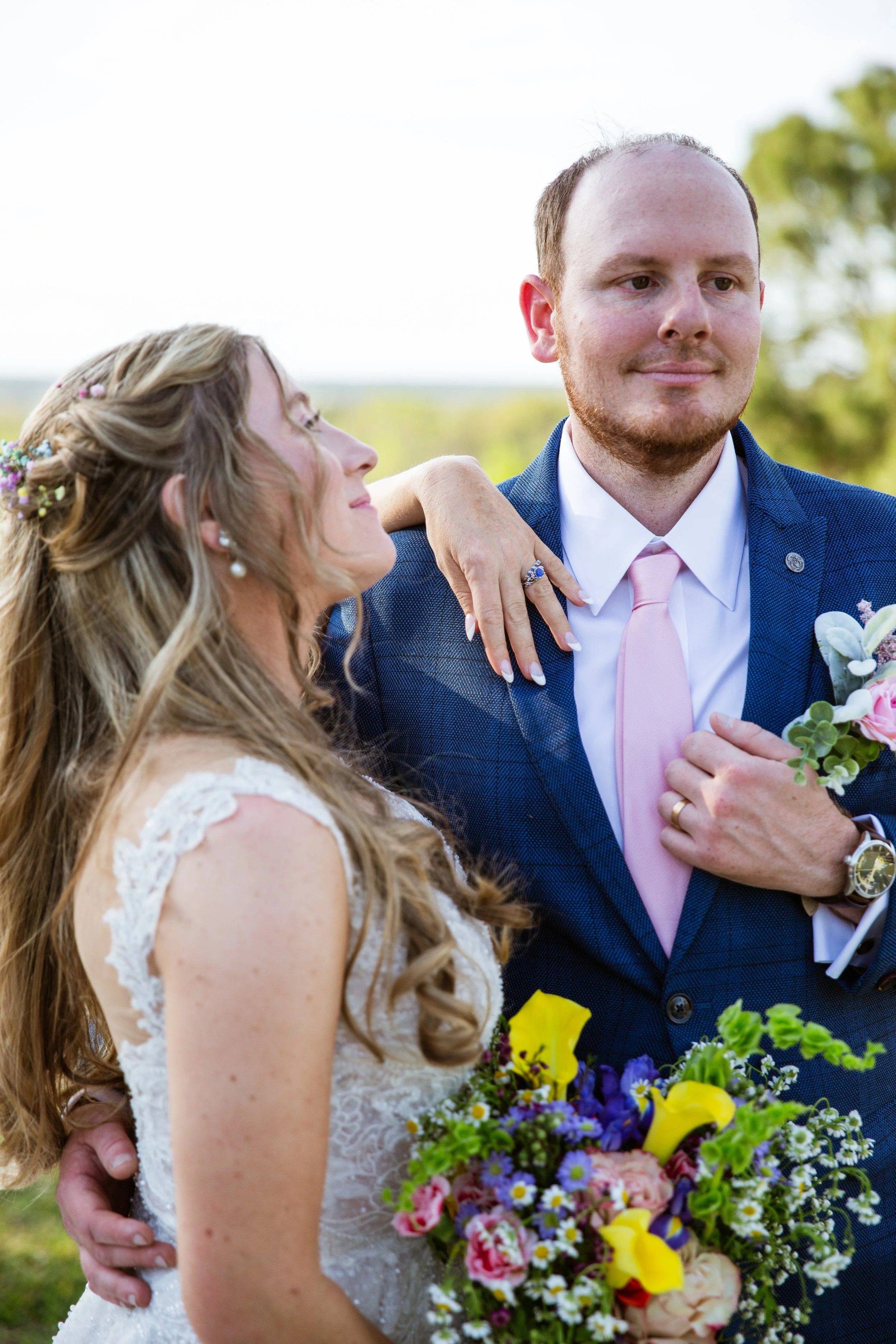 A newlywed couple outside, with the bride holding a colorful bouquet and the groom dressed in a blue suit with a pink tie, embracing and looking at each other with a background of greenery and trees.